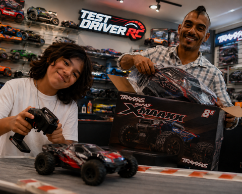A man and a young girl smiling at a hobby shop with radio-controlled cars. The man is holding a large RC car box, and the girl is holding a remote control, with a red RC car on the table in front of her. There are shelves with various RC cars and a sign that says 'Test Driver RC' in the background.