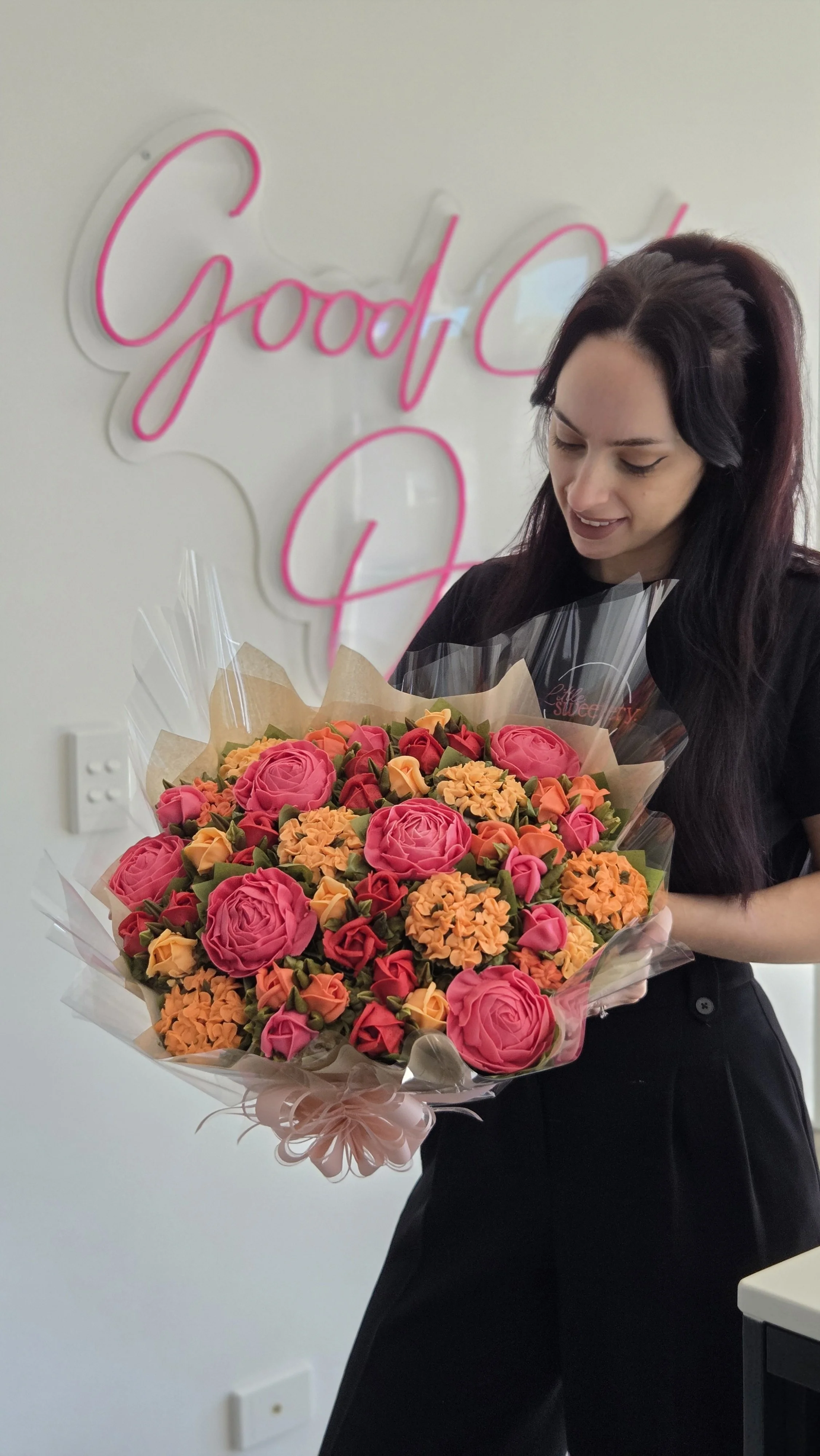 A woman with black and purple hair holding a large bouquet of pink, orange, and yellow flowers in front of a white wall with a neon pink sign that reads 'Good'.