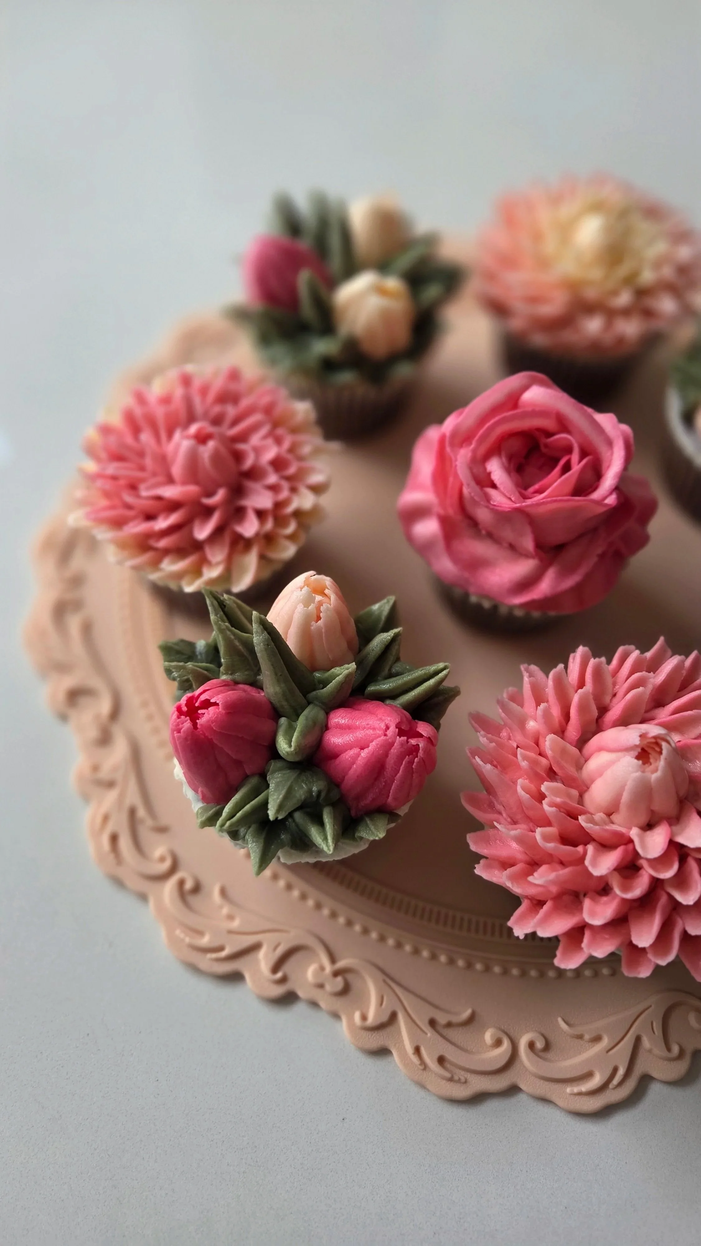Assorted cupcake toppers resembling pink, peach, and cream-colored tulips and chrysanthemums on a decorative pink cake board.