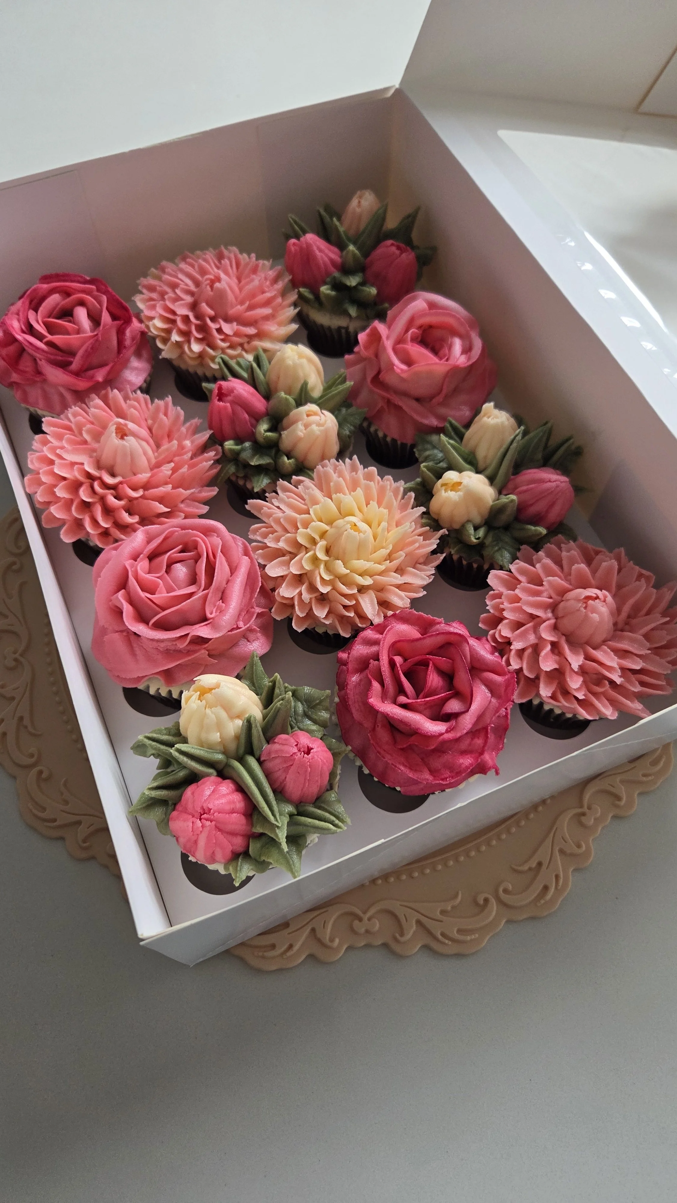 An open white box containing decorative cupcakes with pink and peach floral designs, arranged on a decorative gold tray.