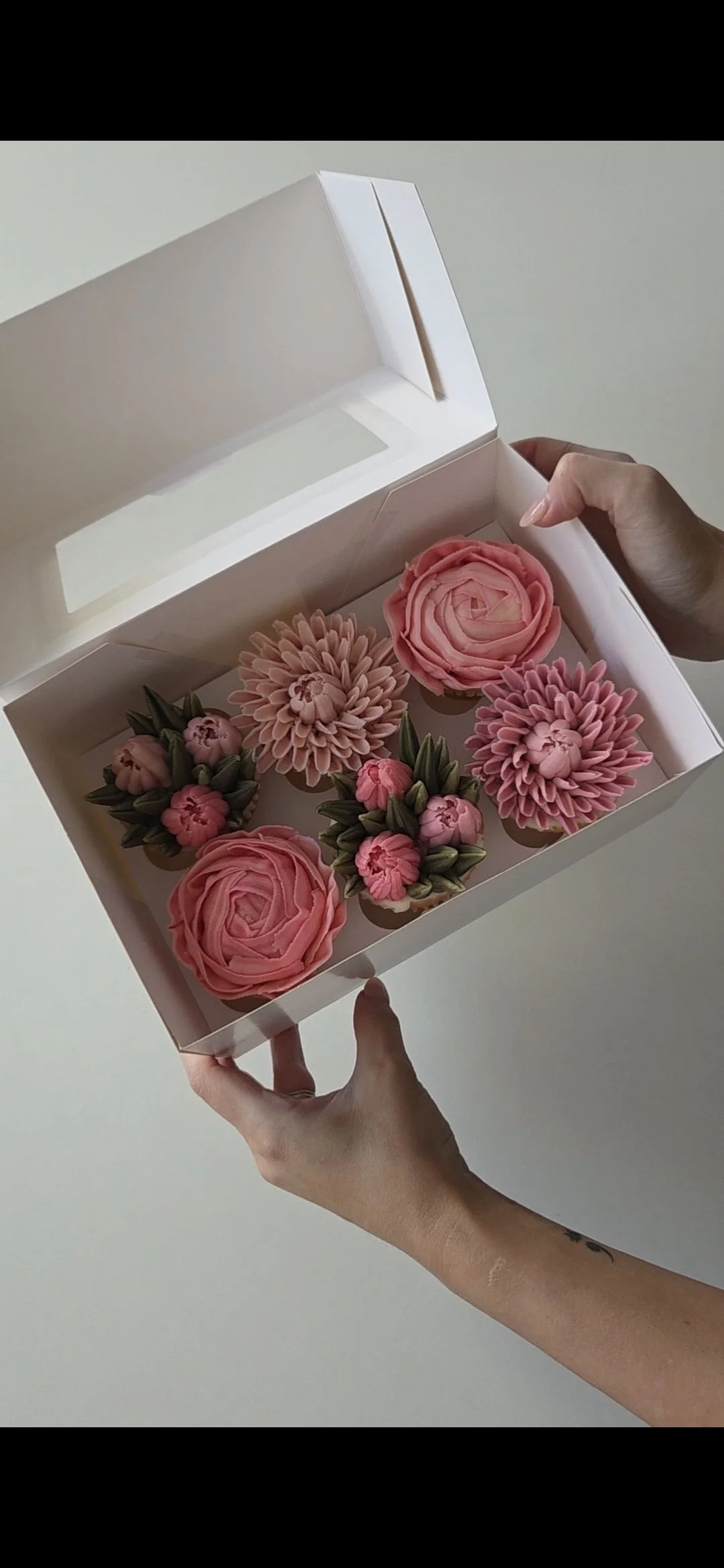 A person holding an open white box with six pink decorative flower cupcakes inside.