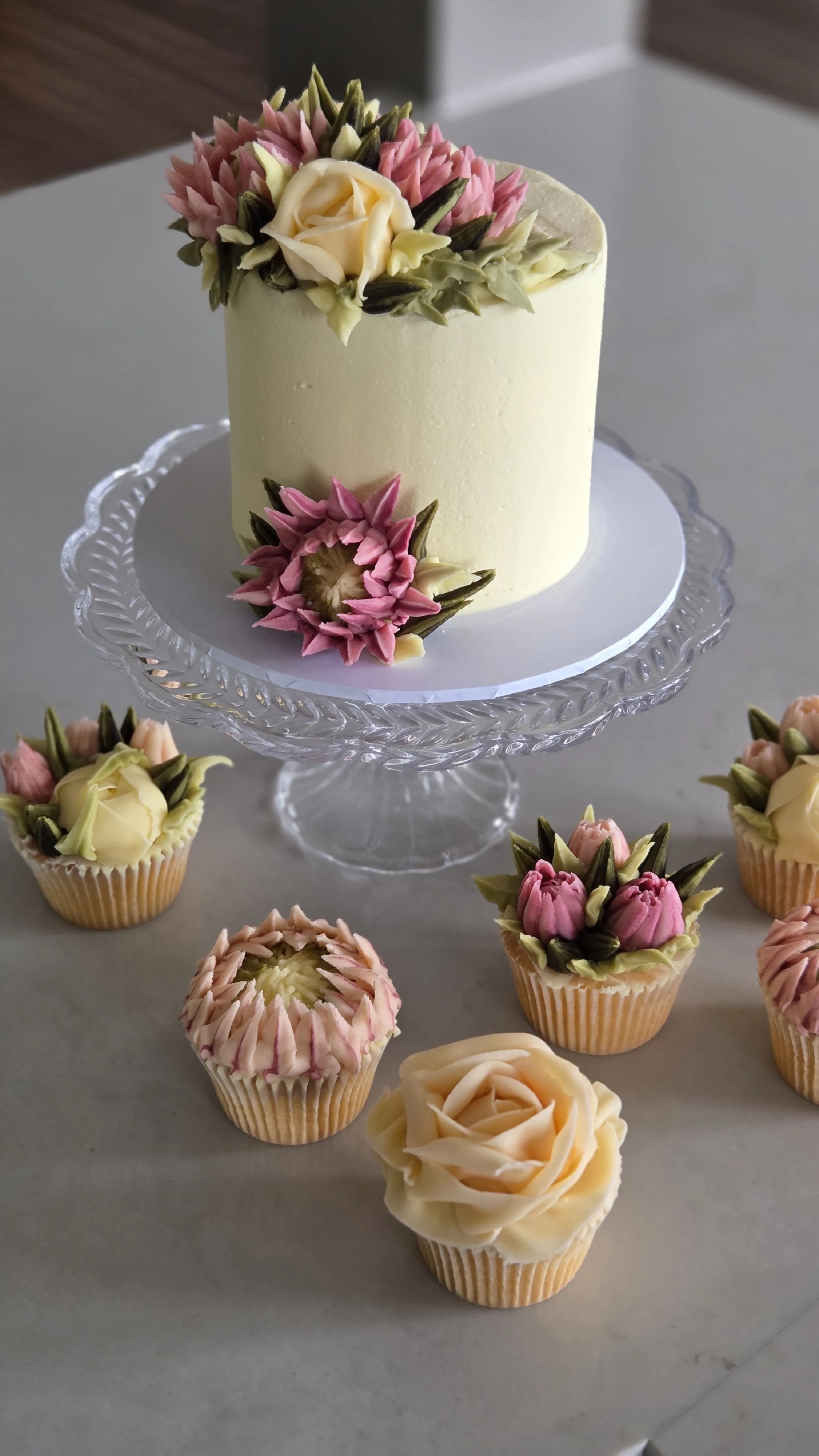 A decorated cake with floral icing designs and several cupcakes with similar floral designs on a glass cake stand, placed on a table.