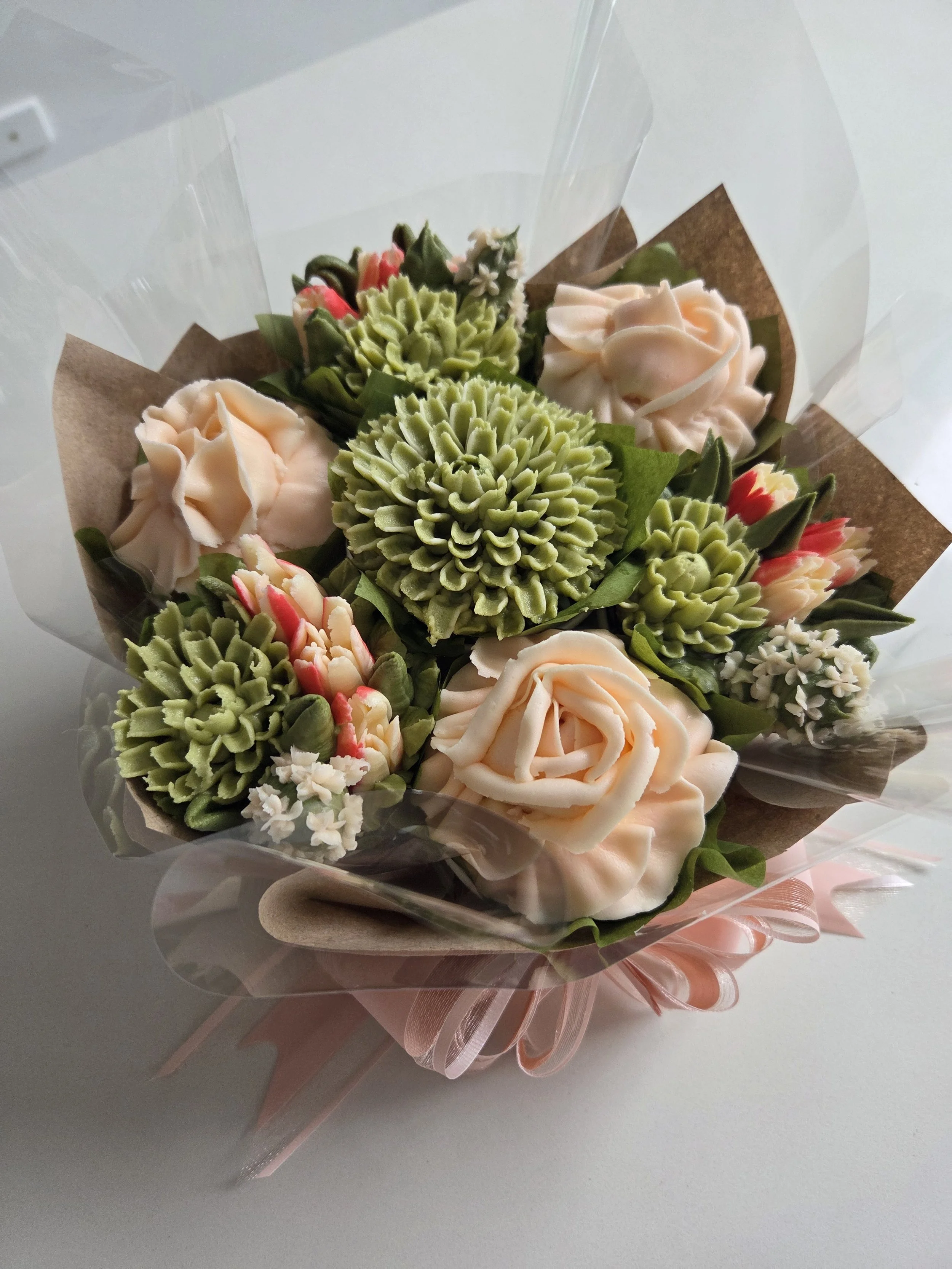 A bouquet of buttercream flowers with light pink roses, green succulents, and small white flowers, wrapped in clear cellophane and brown paper tied with a pink ribbon.