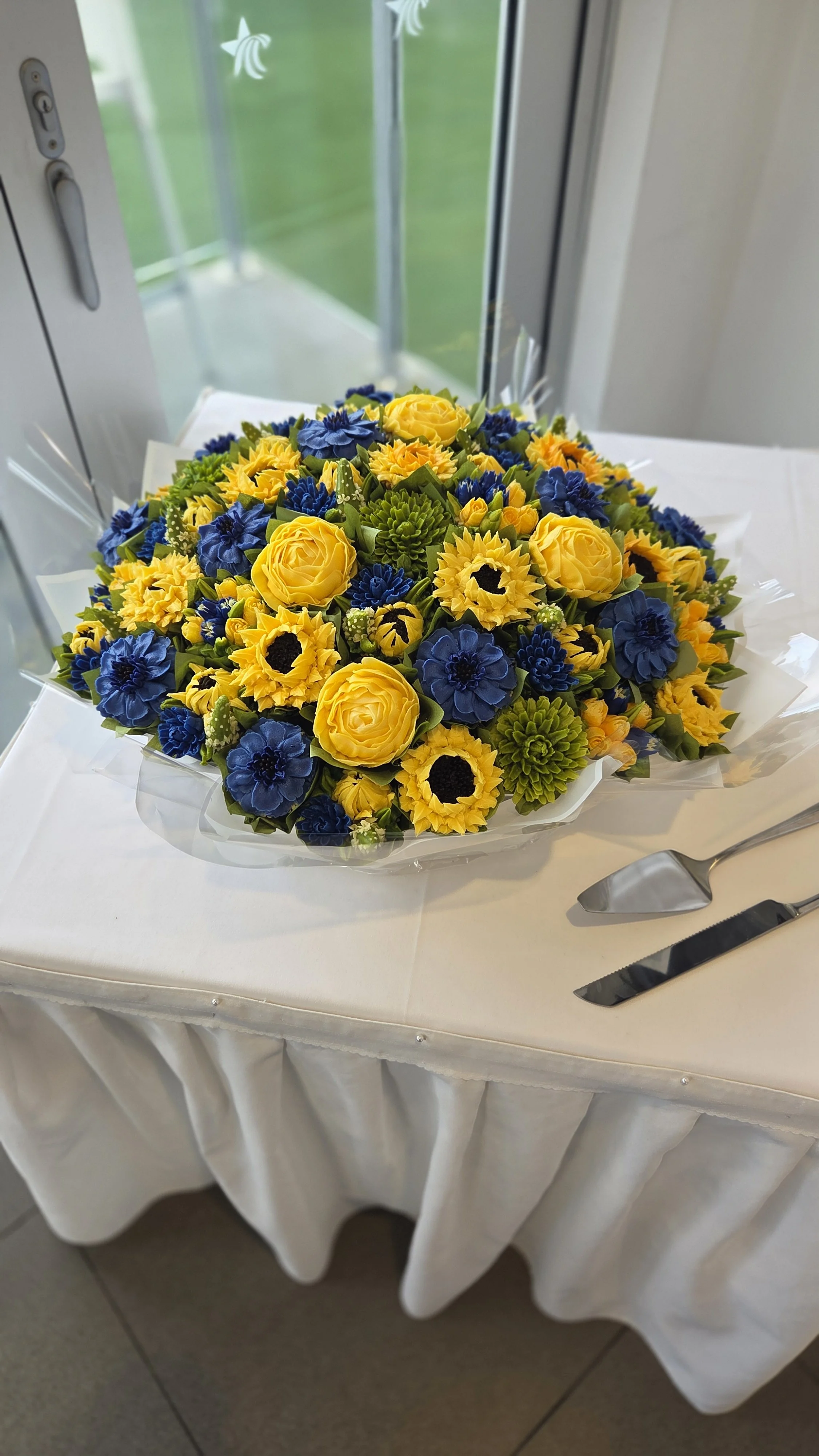A cupcake bouquet of yellow roses, blue flowers, and green foliage on a white table with a cloth cover, next to a cake server and a knife, in front of a window with a green outdoor scene.