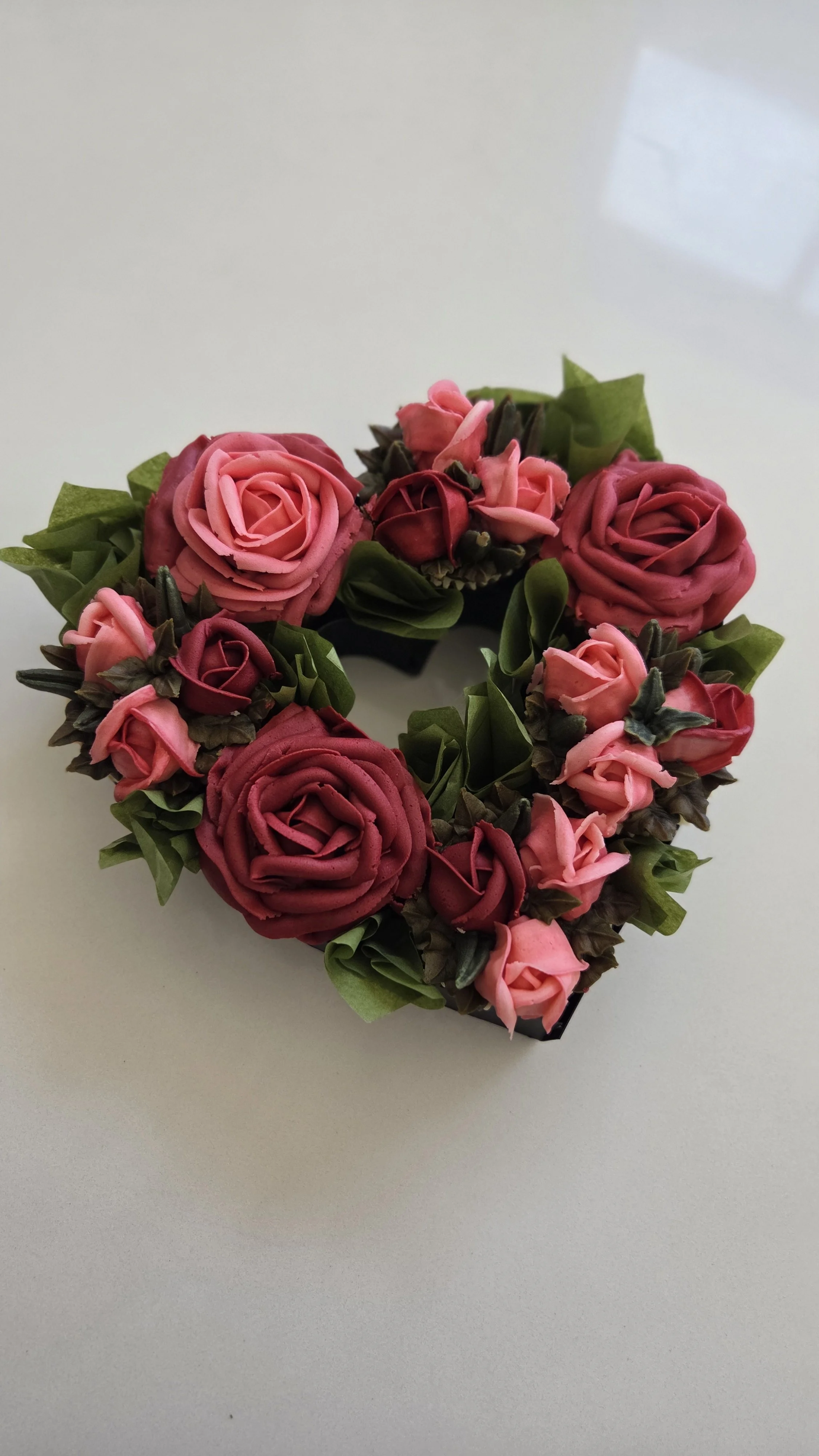 A heart-shaped floral arrangement with pink and burgundy roses and green leaves on a white surface.