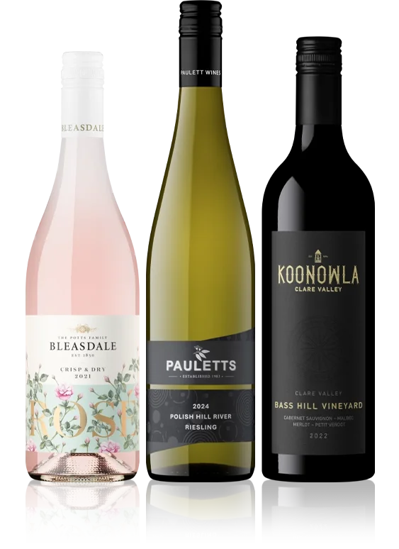 Three wine bottles standing side by side, from left to right: a pink rosé wine bottle with floral label, a yellowish white wine bottle with black and white label, and a dark red wine bottle with black label featuring gold and white text.