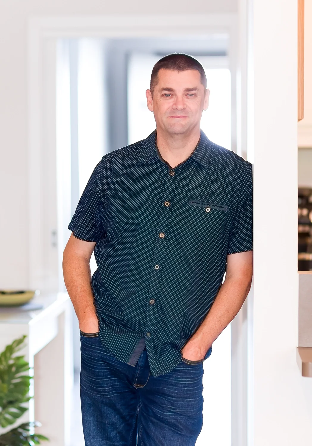 A man with short dark hair, wearing a dark blue short-sleeved button-up shirt with small white dots, standing in a well-lit room with his hands in his pockets, leaning against a doorway.