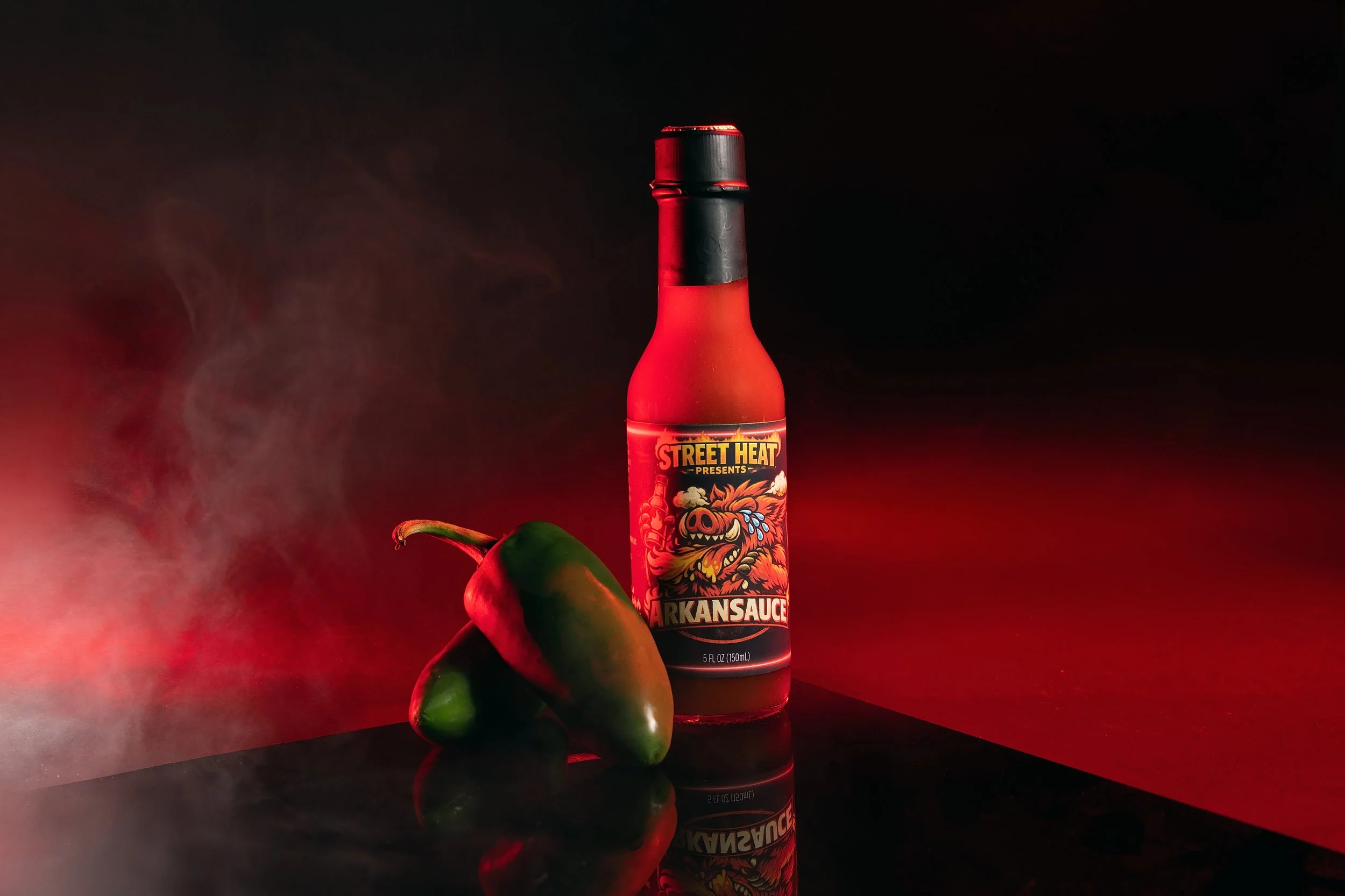 A bottle labeled 'Arkansas Sauce' with a dragon illustration on it, accompanied by two green jalapeño peppers against a dark background with red lighting and smoke.