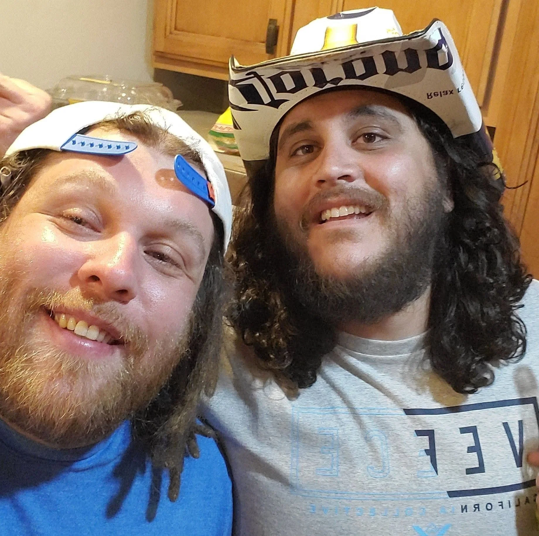 Close-up of two men smiling, one with a backward white baseball cap and a blue strap, the other with a newspaper hat and dreadlocks, indoors with wooden cabinets and a countertop in the background.