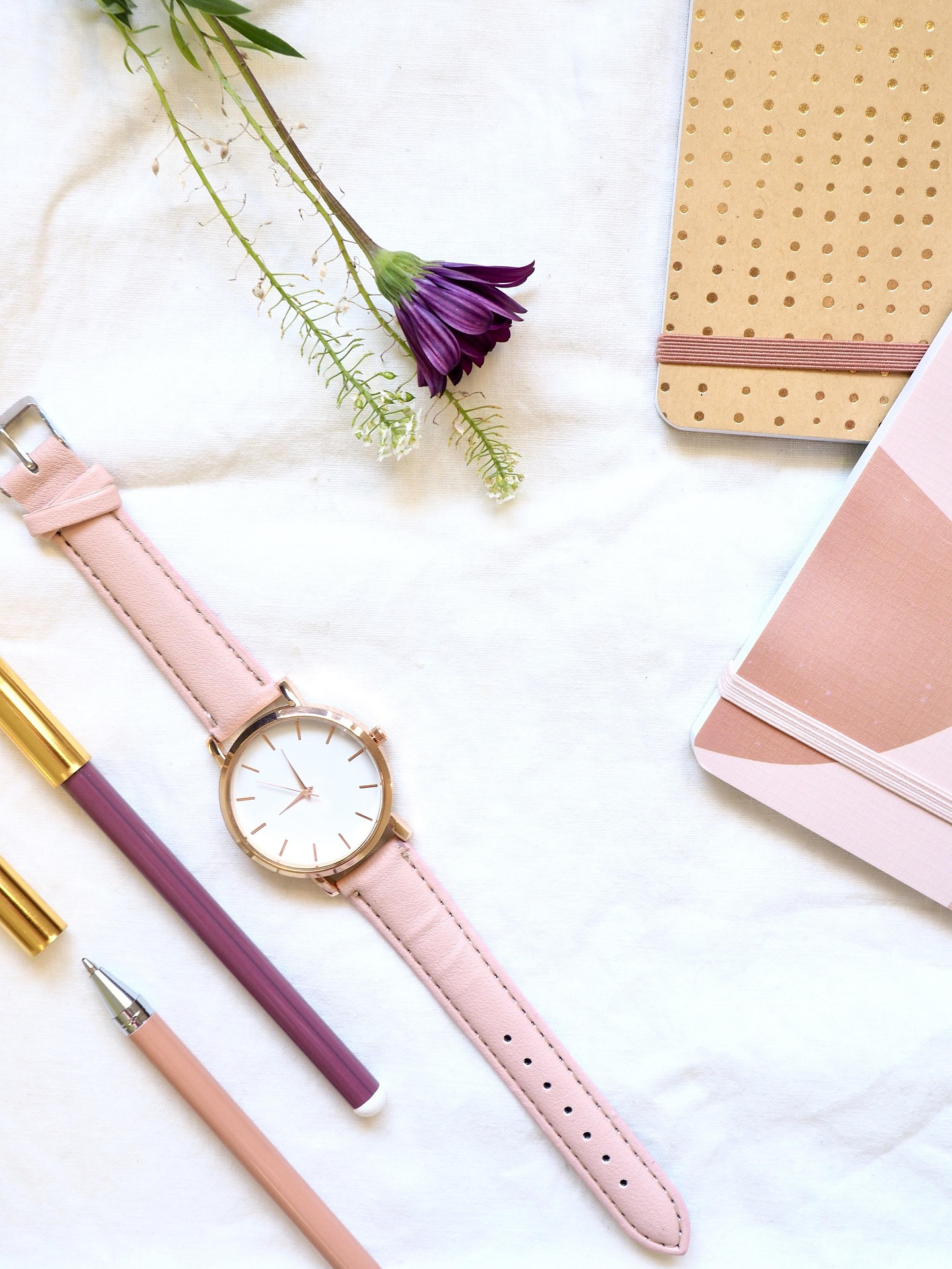 A flat lay of a pink wristwatch, purple and pink pens, a pink notebook, a floral branch, and a beige notebook on a white surface.