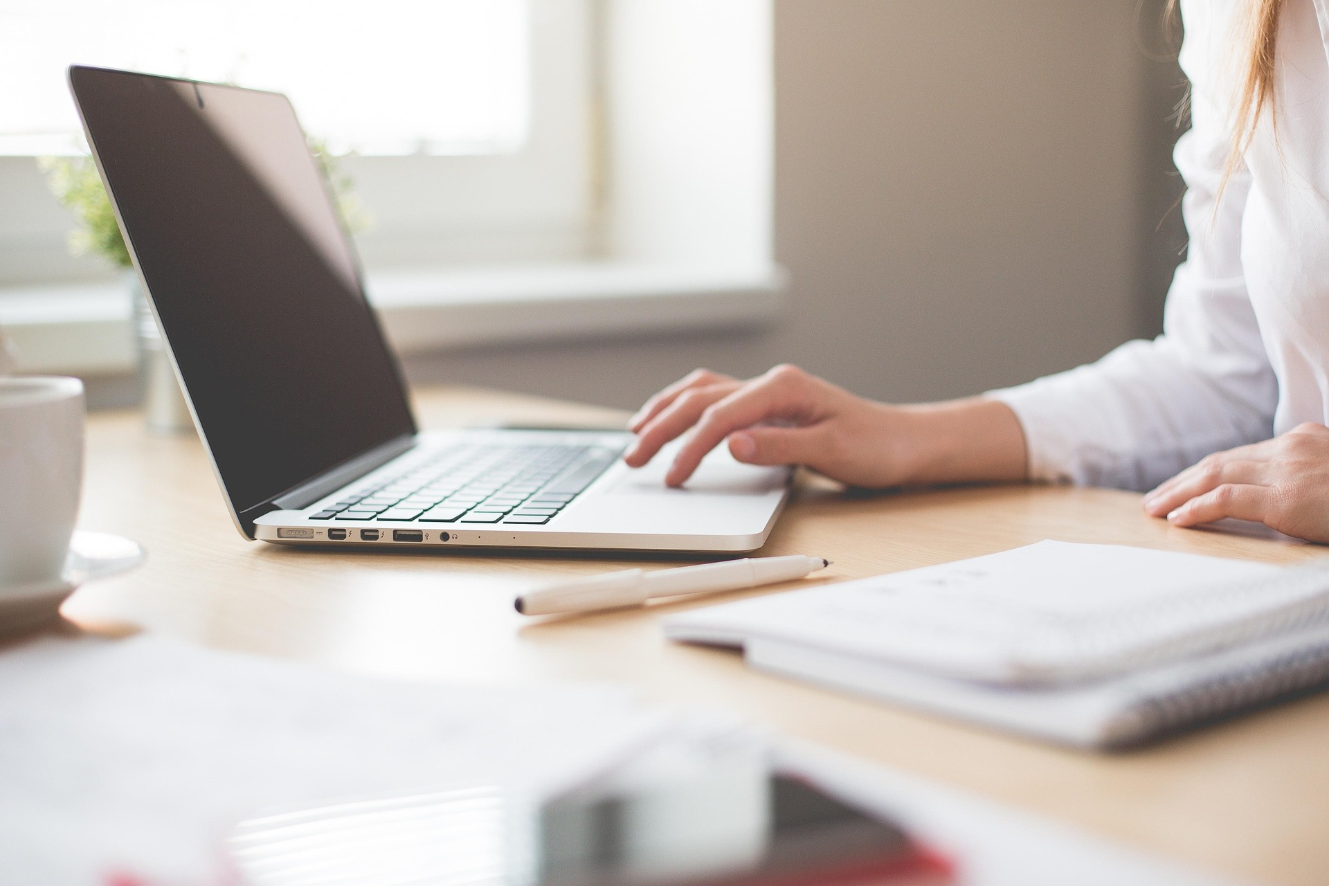 Person using a silver laptop on a wooden desk with papers, a pen, a potted plant, and a coffee mug nearby.