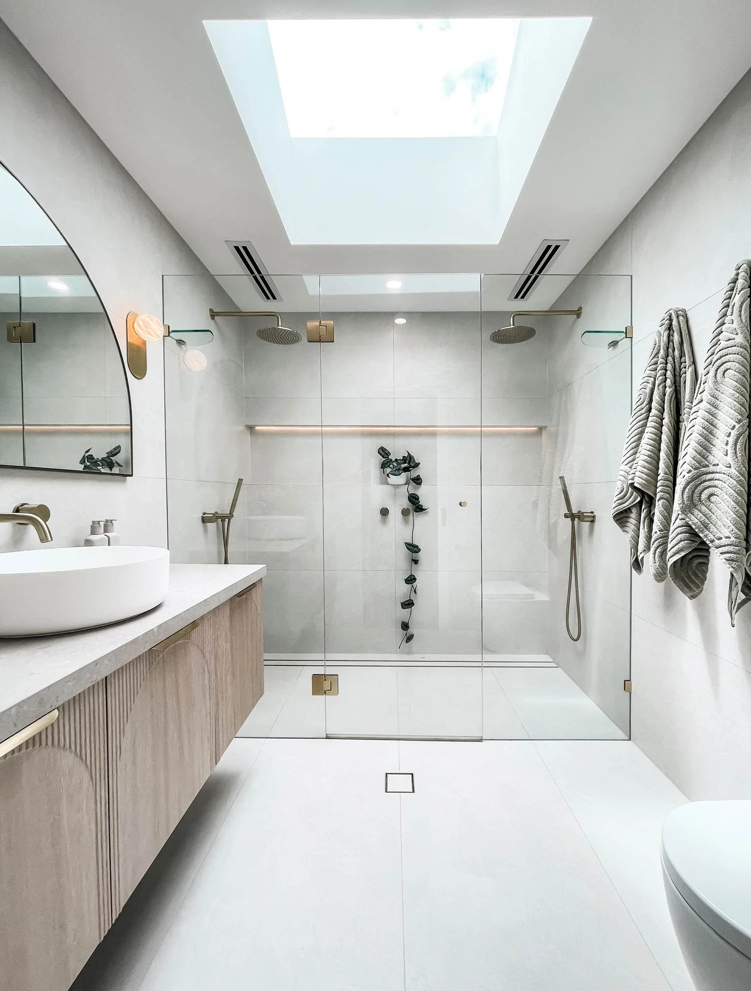 Modern bathroom with a skylight, tiled walls, a wooden vanity with a vessel sink, a large mirror, and a glass-enclosed shower with two rainfall showerheads.