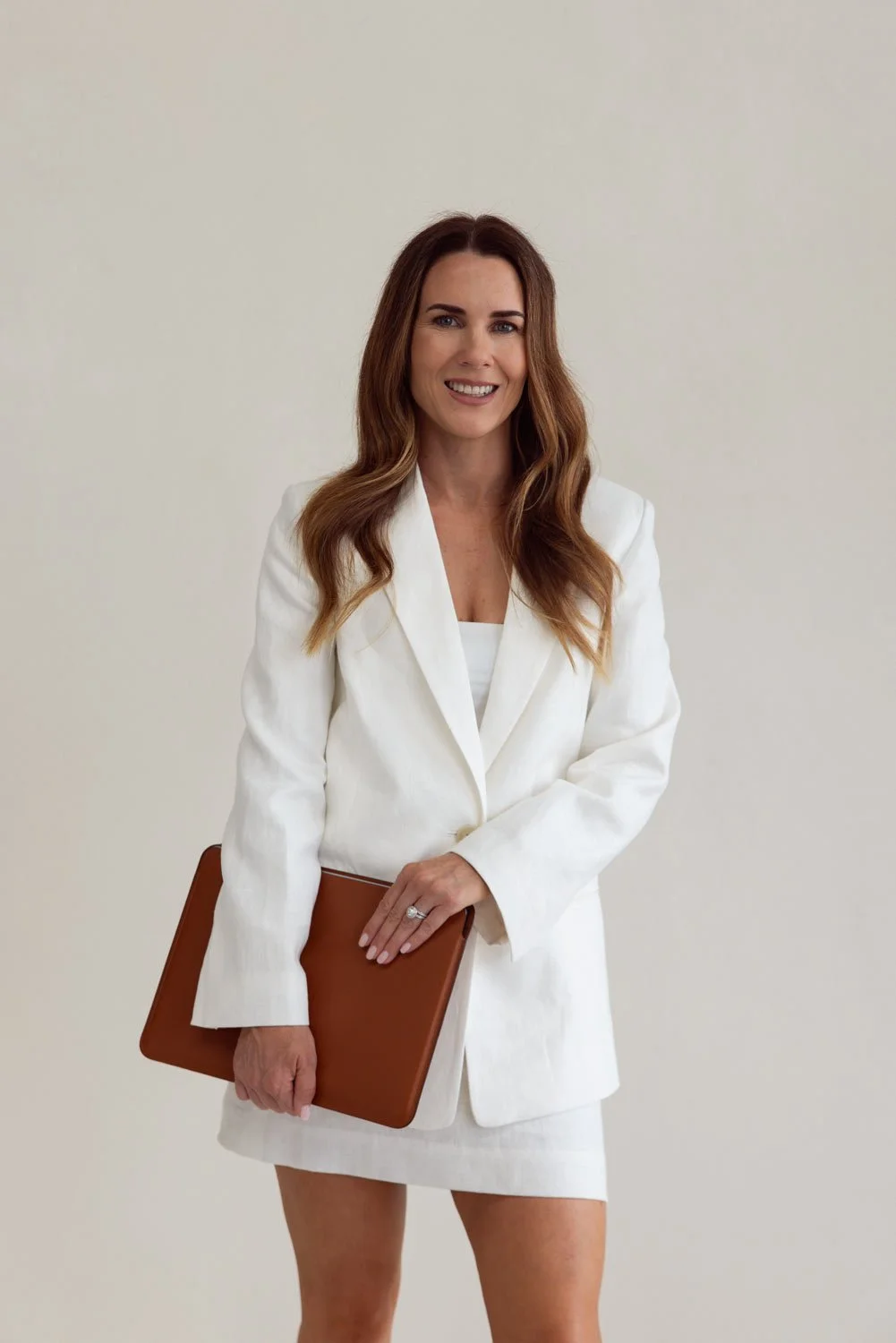 Gold Coast residential interior designer, Christine Fletcher, in a white blazer and skirt, holding a brown folder and smiling, standing against a plain white wall.