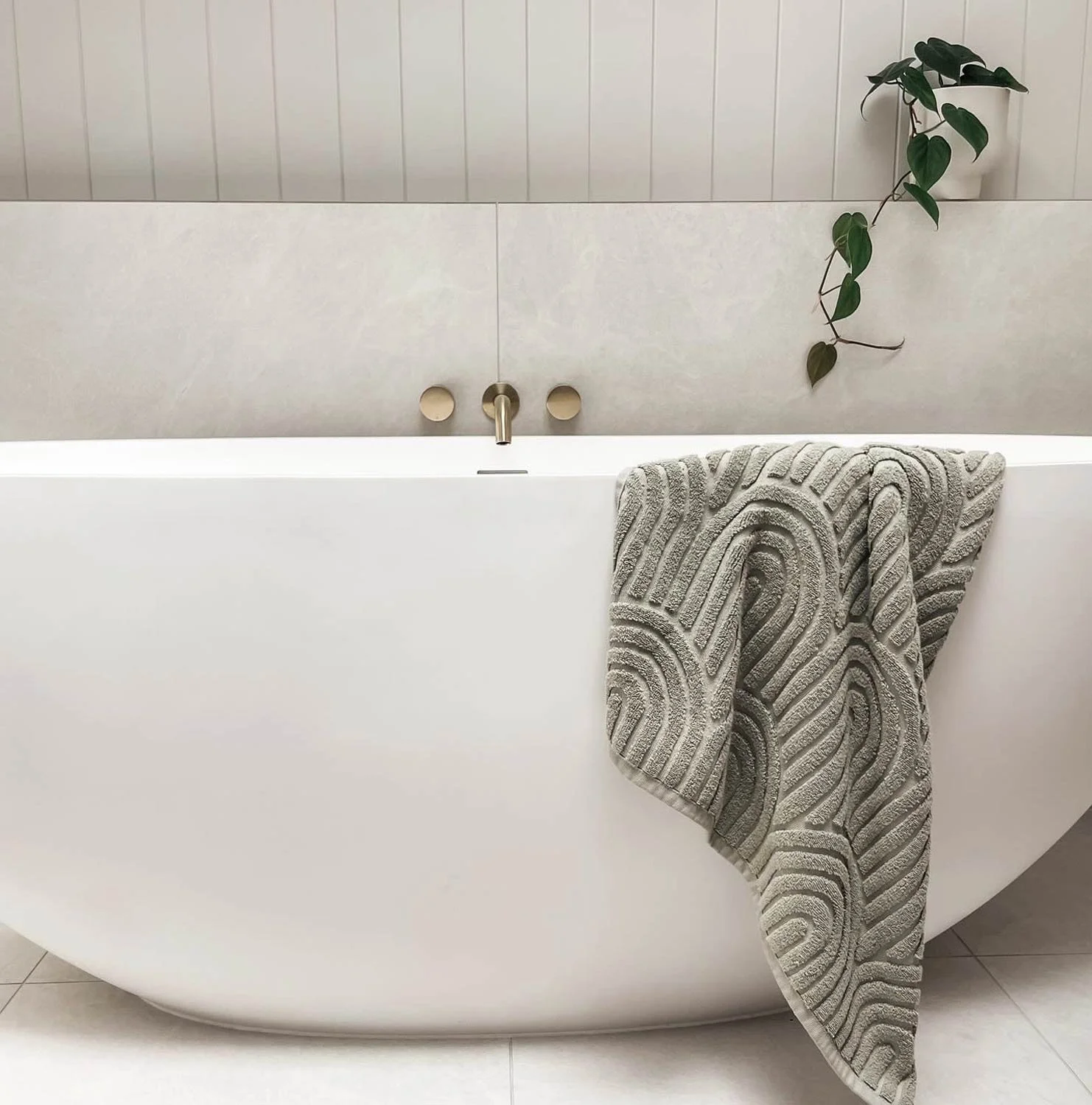 Freestanding bath with green towel hanging over edge, plant in pot on shelf, and wooden wall paneling behind.