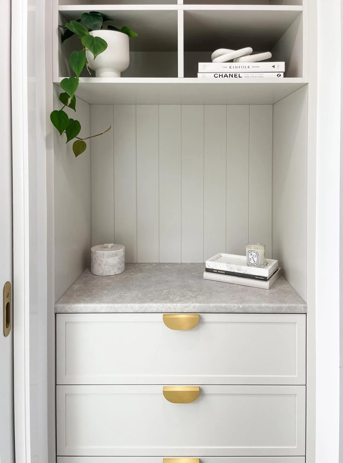 Interior of a white built-in cabinet with gold handles, containing decorative items like a potted plant, books, and candles.