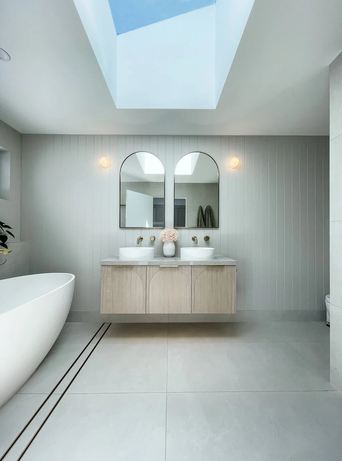 Modern bathroom with a freestanding bathtub on the left, a double vanity with two sinks, two arched mirrors, and a skylight ceiling on top.