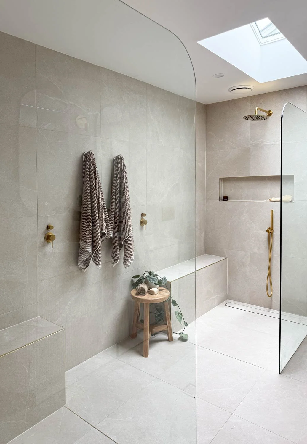 A modern, minimalist walk-in shower with beige marble tiles Two towels hang on hooks on the left wall, and a small wooden stool holds a rolled towel, soap, and a plant. The shower has a gold showerhead and a rain shower fixture, with a glass partitio