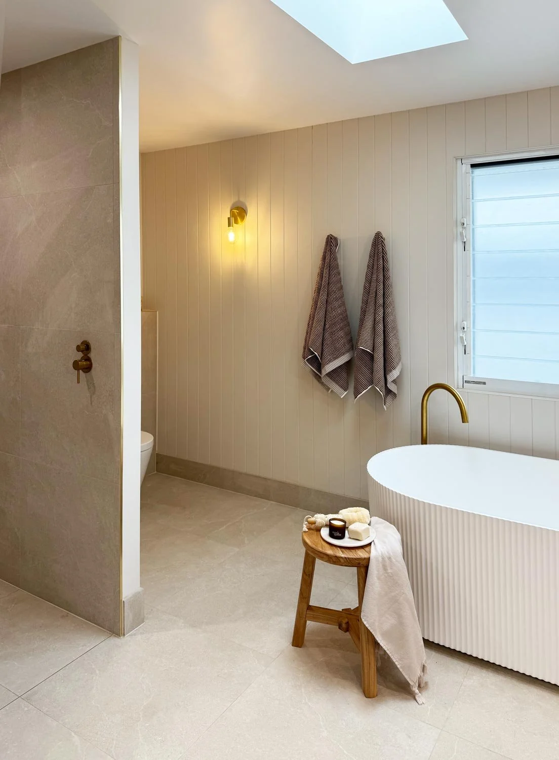 Modern bathroom with bathtub, two bath towels hanging on the wall, a window, and a small wooden stool with toiletries.