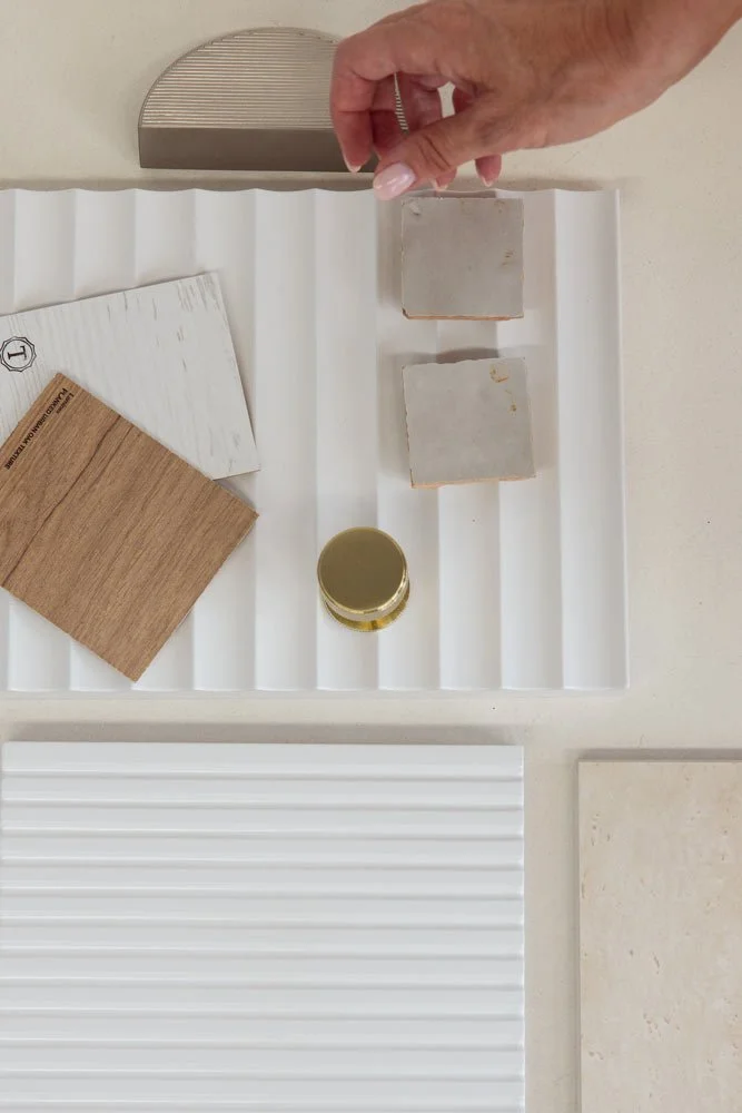A hand placing a small tile on a display tray with various tile samples, including wood grain, marble, and ceramic tiles.