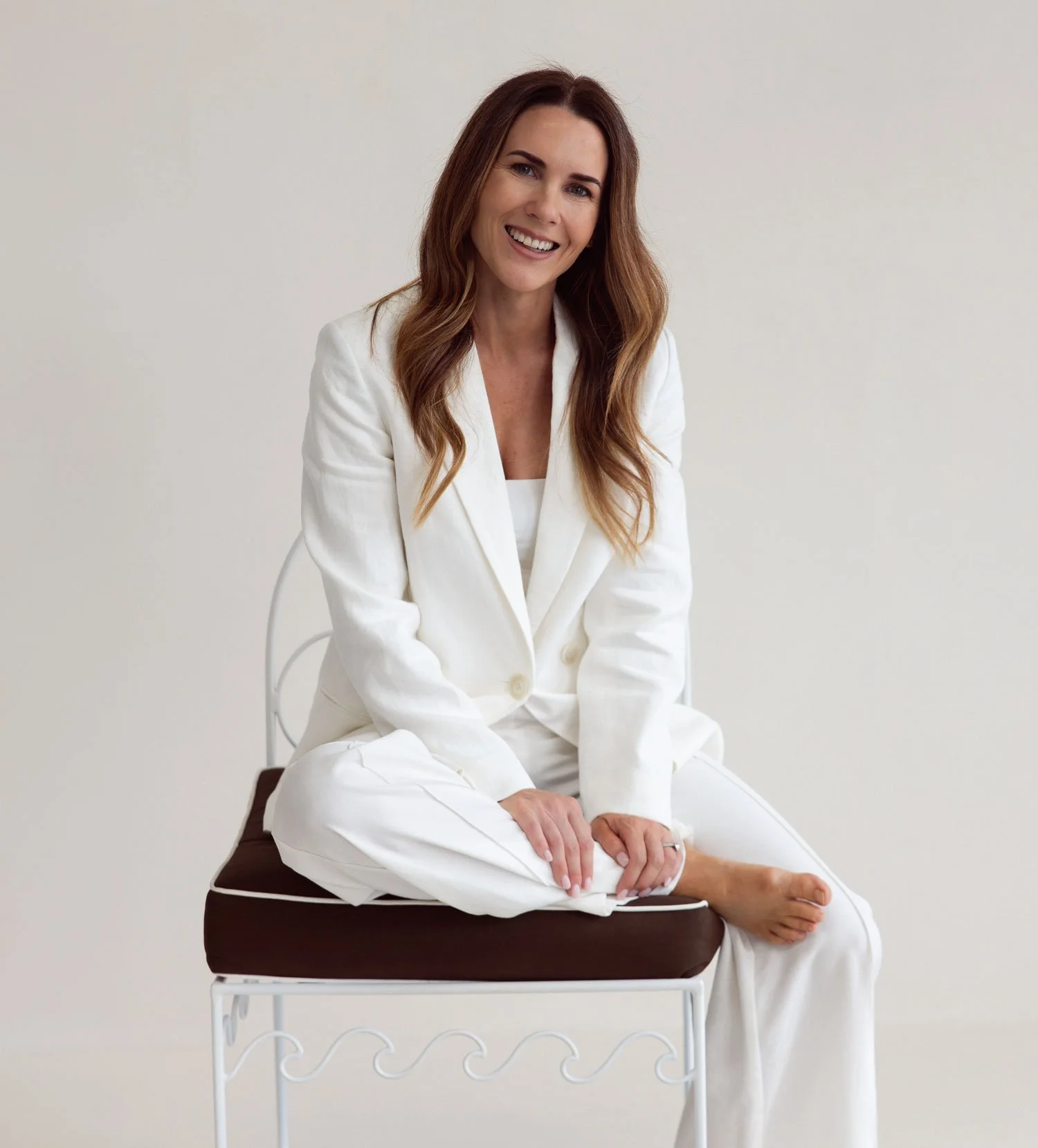 Christine Fletcher, Queensland interior designer, sitting on a brown cushioned metal framed chair, wearing an all-white suit, smiling against a plain light background.