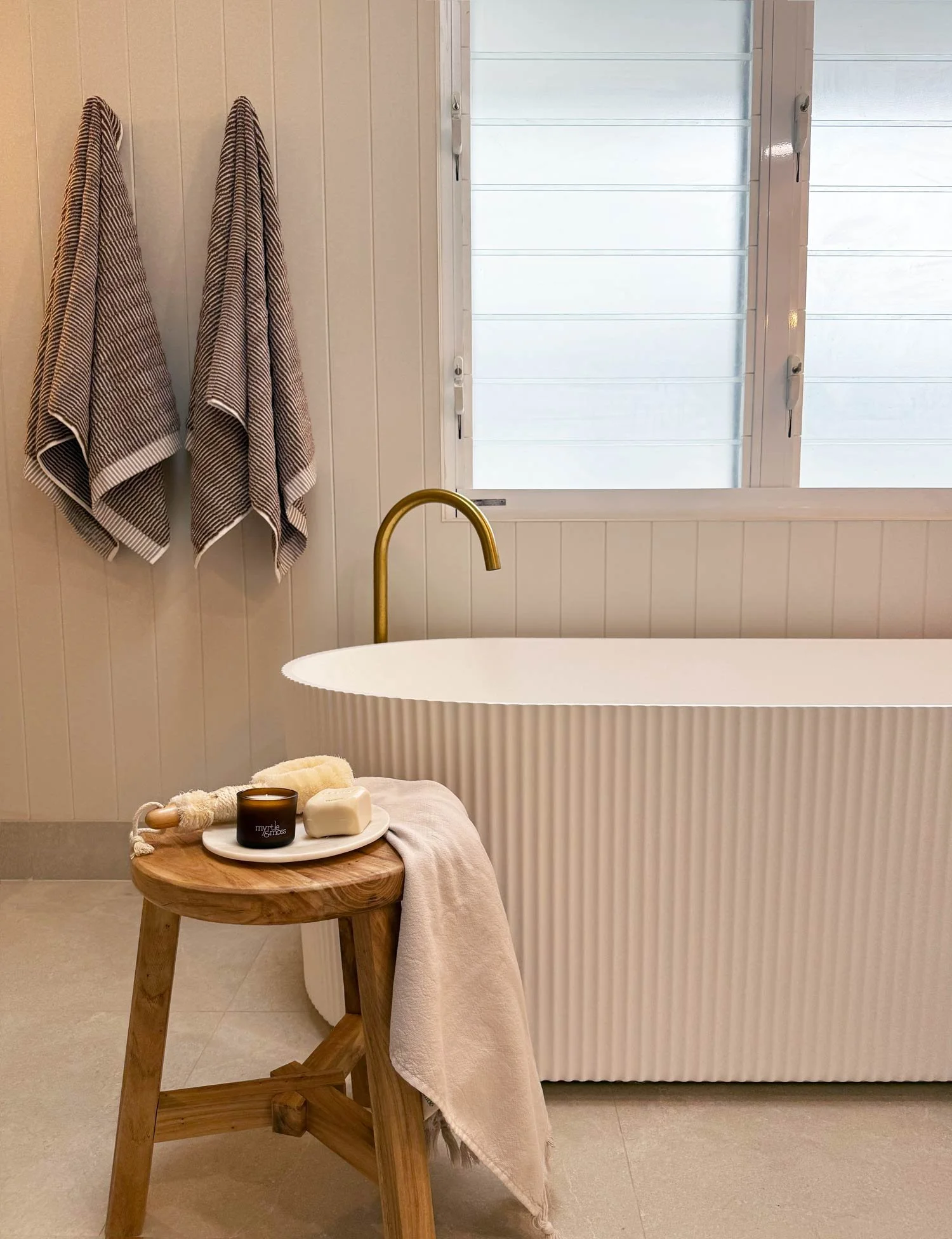 Freestanding bath with two towels hanging on wall and wooden stool in front with candle, soap and towel.