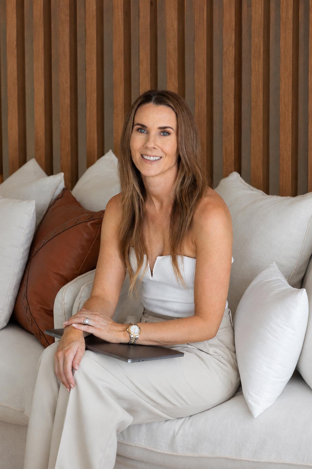 Christine Fletcher, Queensland interior designer, sitting on a beige sofa with white and brown pillows, smiling at the camera, with a wooden panel wall in the background.