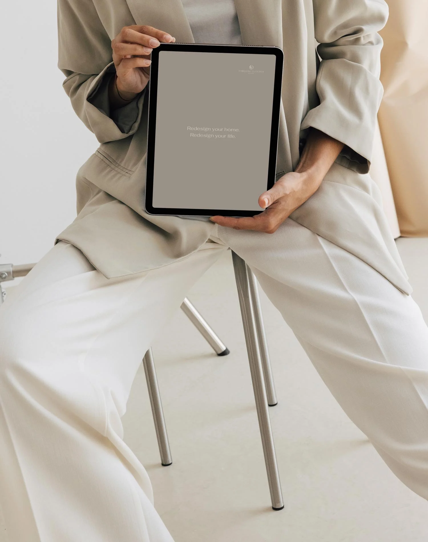 Person sitting on a stool holding a tablet with a neutral-colored screen that has text 'Redesign your home. Redesign your life.'.
