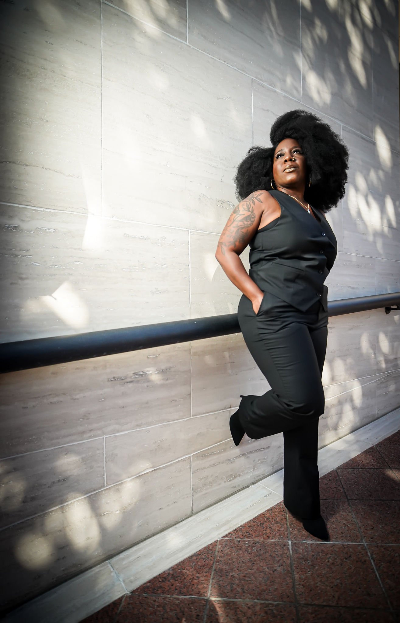 A Black woman with large curly hair, wearing a black sleeveless top and black pants, posing against a wall with shadows of leaves, hands in pockets, one foot resting on a ledge.