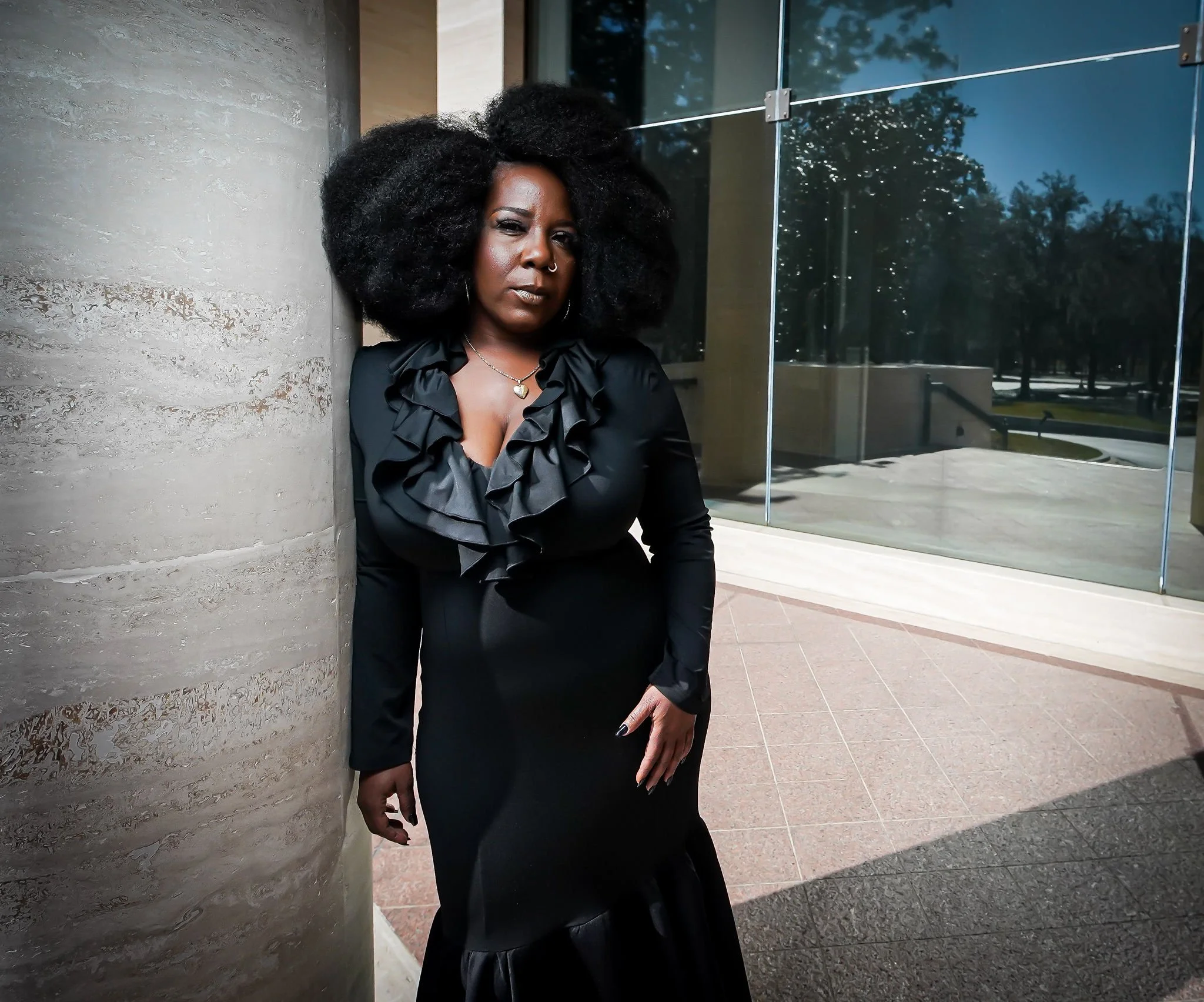 A woman with large afro hairstyle wearing a black dress with ruffled neckline, accessorized with a necklace and nose piercing, standing outdoors near a modern building with reflective glass and stone wall.