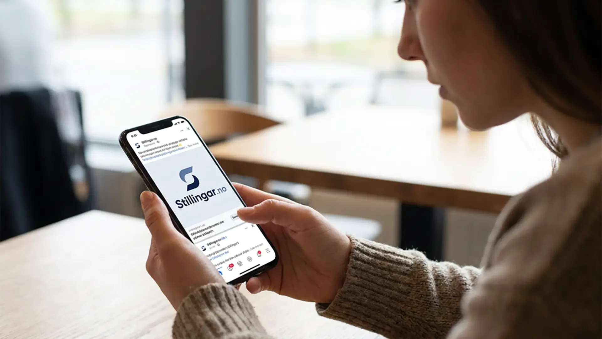 Person ser på mobilen sin med Stillingar.no-logo på skjermen, sittende i et rom med store vinduer.
