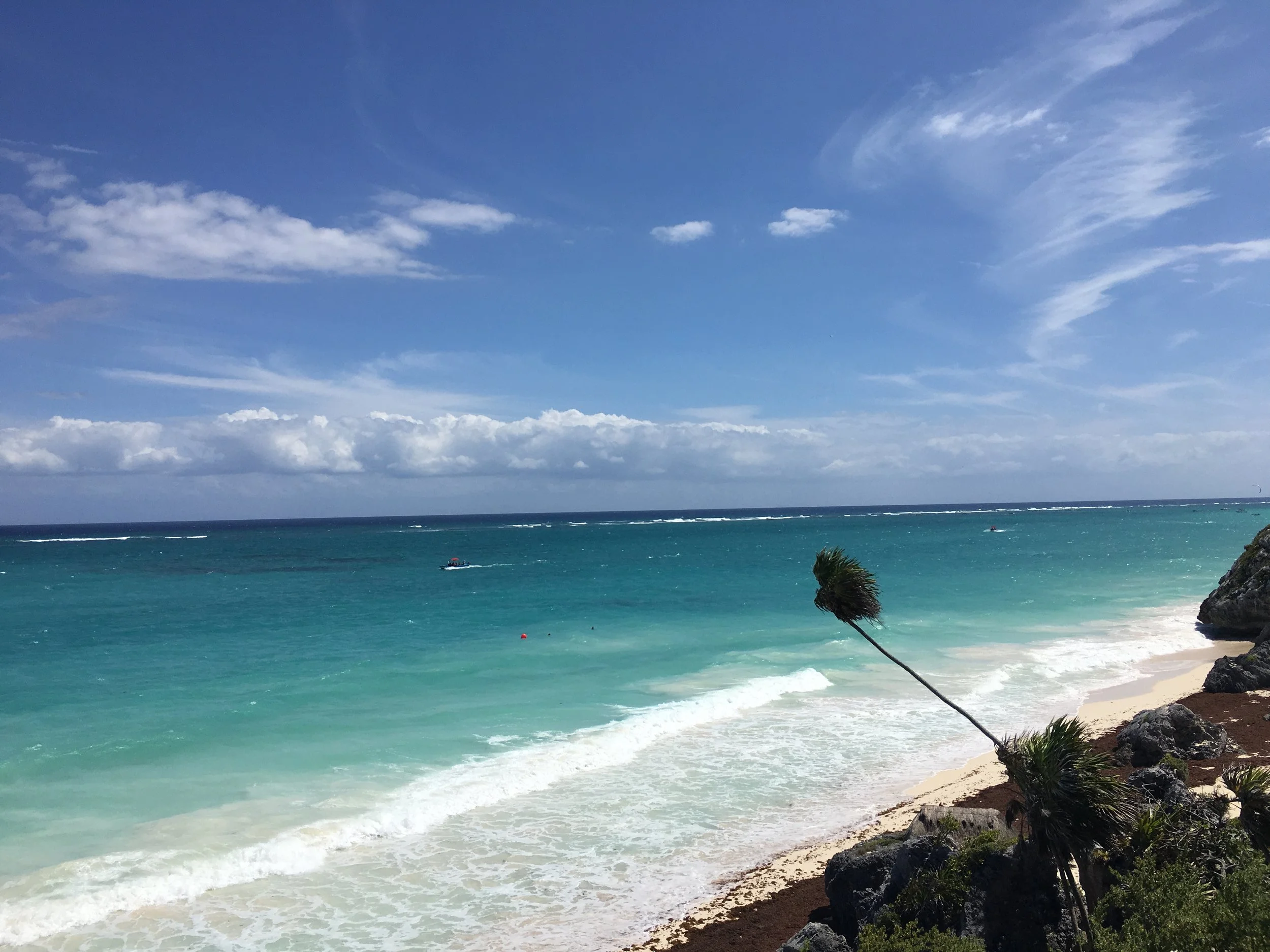 A beach with turquoise water and white sand, rocks and palm trees in the foreground, and a partly cloudy sky overhead.