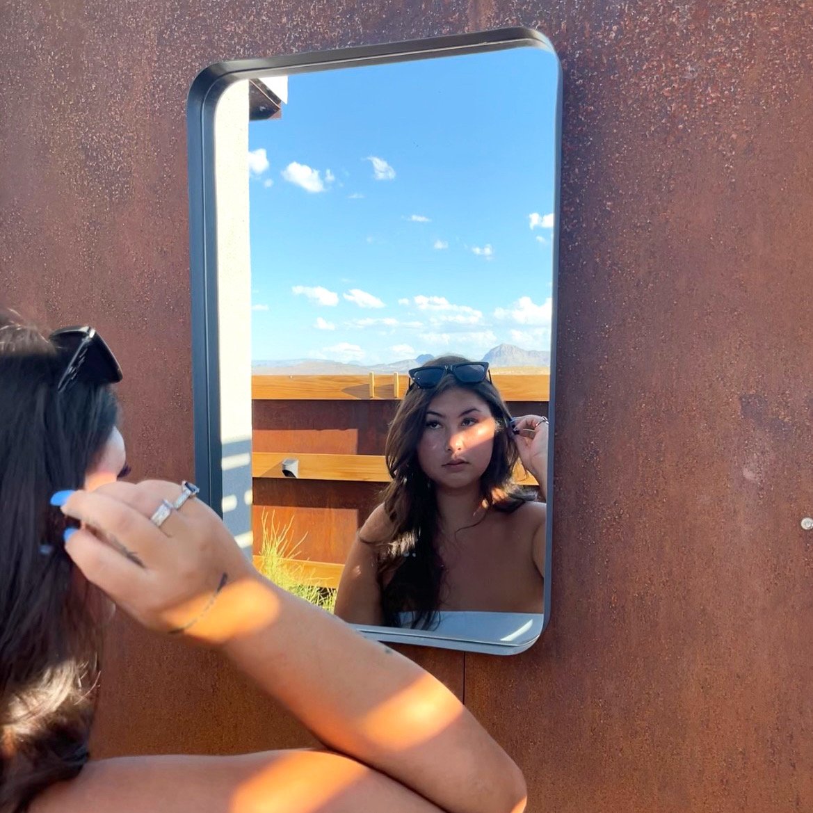A woman is looking at her reflection in a mirror mounted on a rusty-colored wall outdoors. She is adjusting sunglasses on her head, with a clear blue sky and distant mountains in the background.