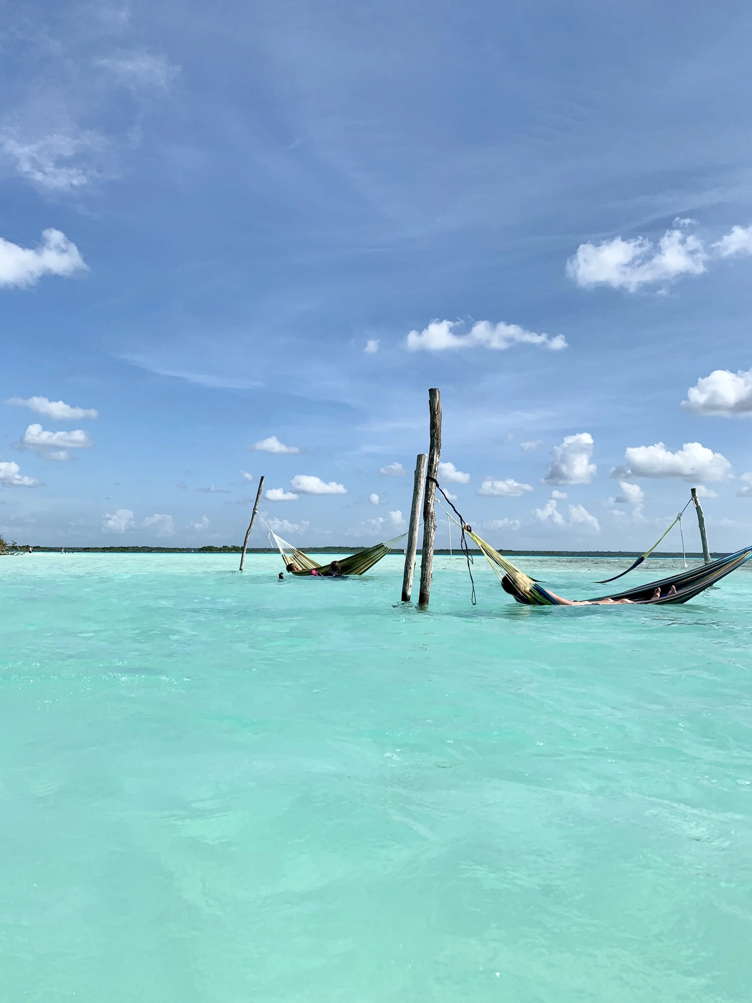 A tropical beach scene with turquoise water, wooden posts, and a person relaxing in a hammock tied between two posts under a blue sky with scattered clouds.