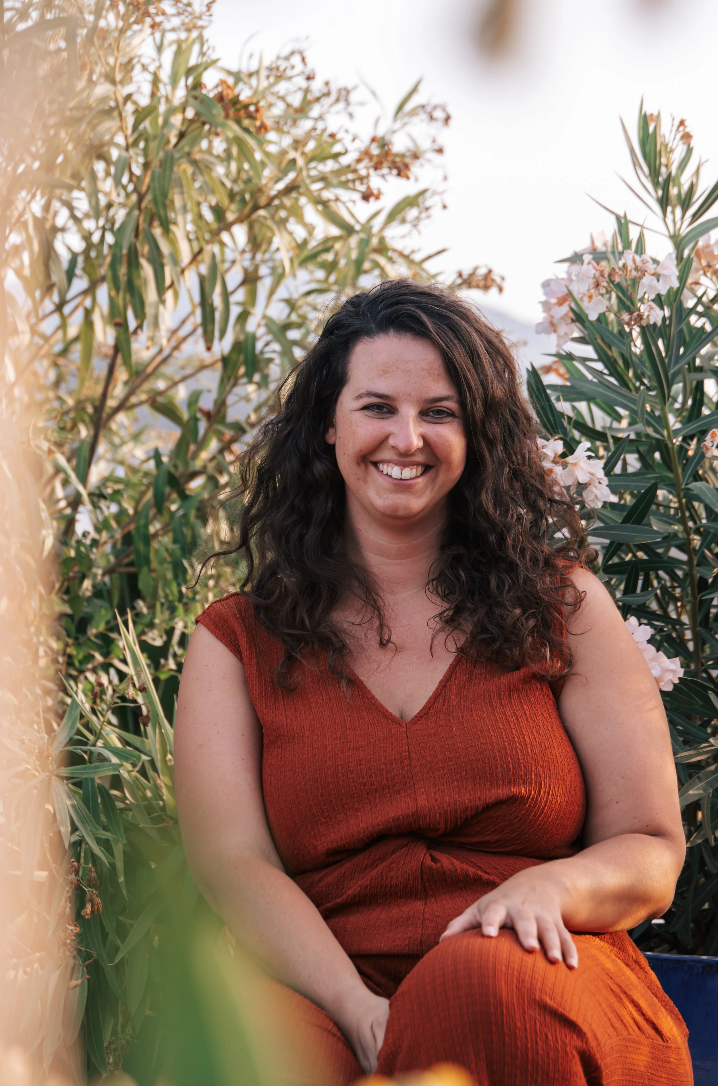 A woman with curly hair smiling and sitting outdoors among green plants and pink flowers, wearing a sleeveless orange dress.