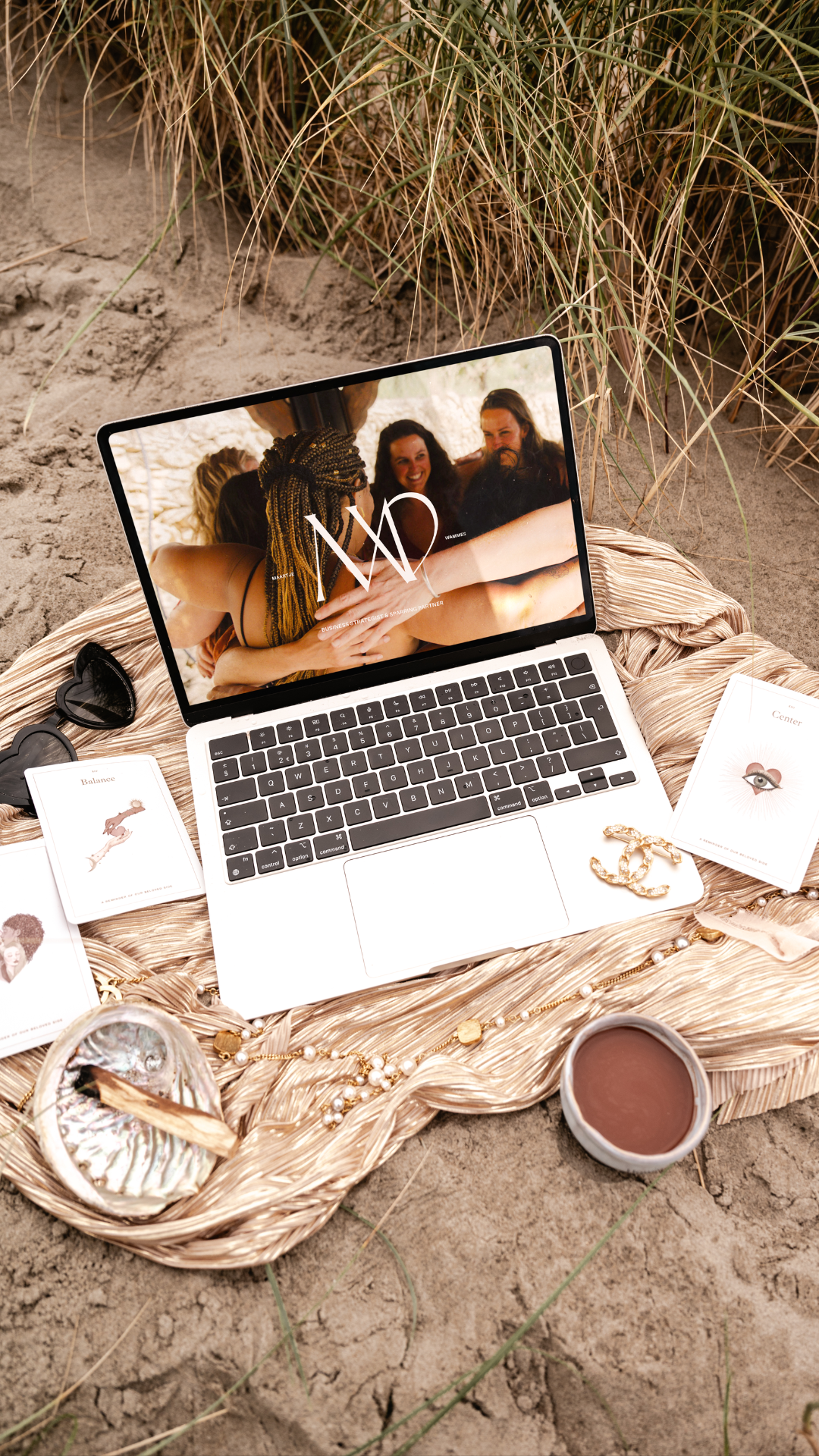 Laptop on a beach showing the Maartje Wammes homepage, "Business Strategist & Sparring Partner," surrounded by oracle cards, cacao and jewelry
