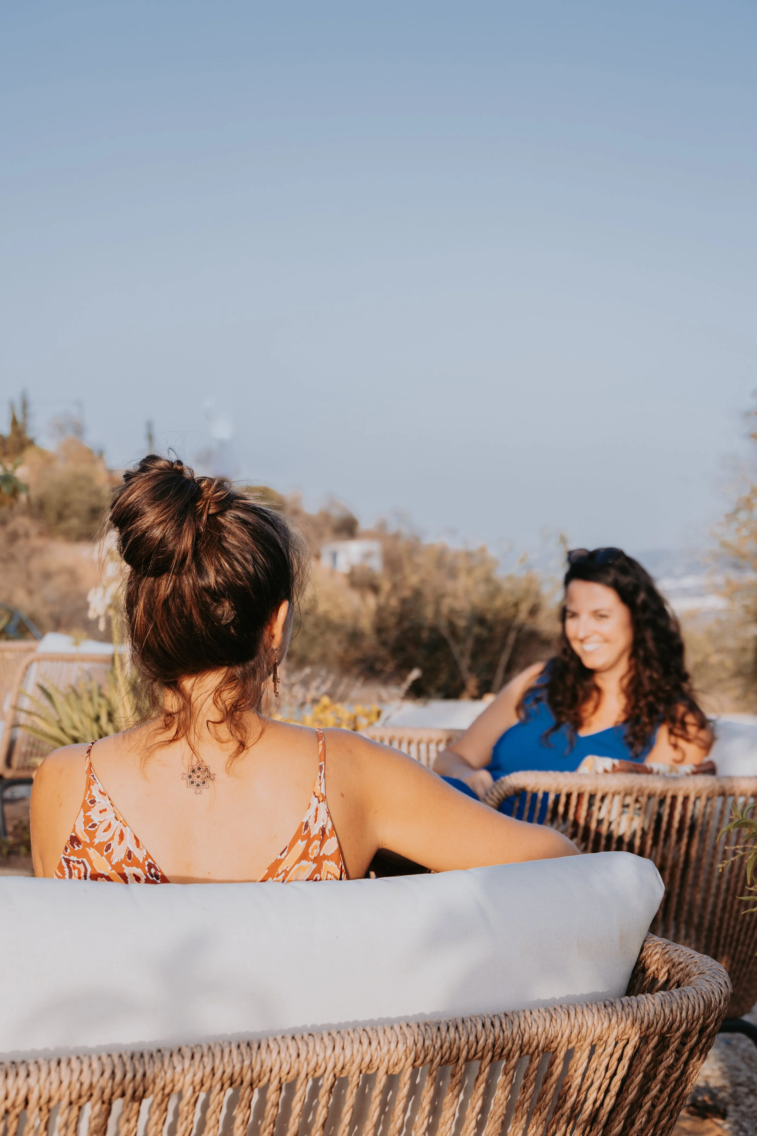 Two women sitting outdoors on wicker chairs, engaging in conversation, with a woman in the foreground having her back to the camera. The setting appears to be a patio or garden area with trees and distant hills under a clear sky.