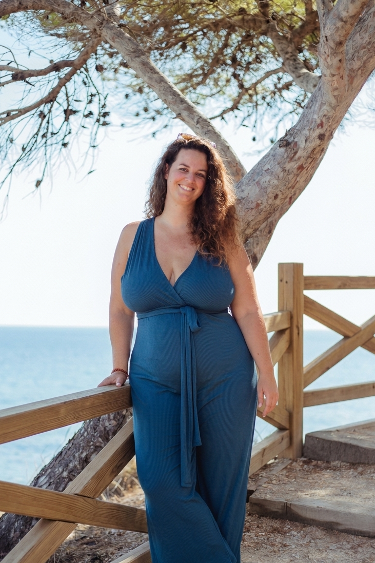 A woman in a blue sleeveless jumpsuit leaning on a wooden railing near the beach, smiling, with a large tree and the ocean in the background.