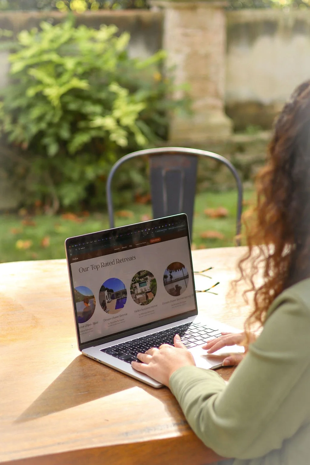 A woman with curly hair using a laptop outdoors on a wooden table, with greenery and a stone wall in the background.