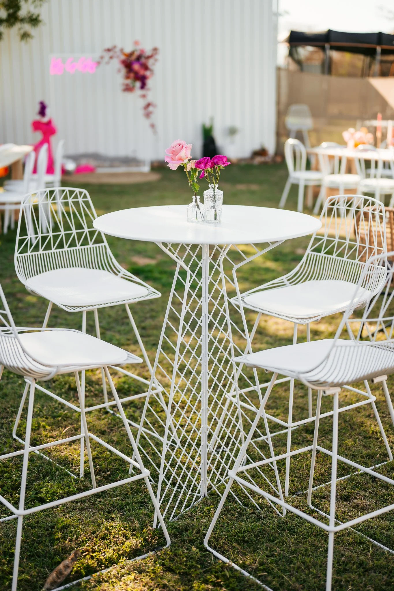 White outdoor table with decorative metal chairs and pink roses in glass vases, set in a garden with other tables and chairs in the background.