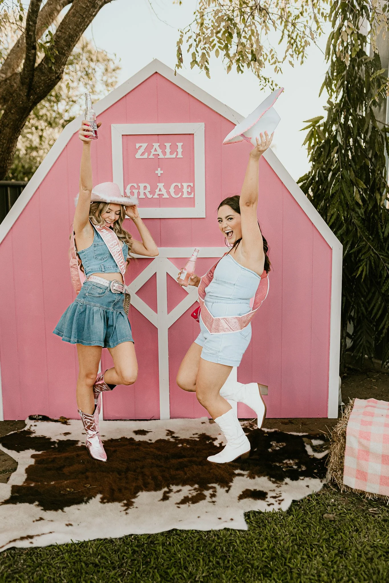 Two women smiling and dancing in front of a pink barn with the sign 'Zali + Grace,' holding bottles and wearing fashionable summer outfits with cowboy boots, surrounded by trees and outdoor decorations.