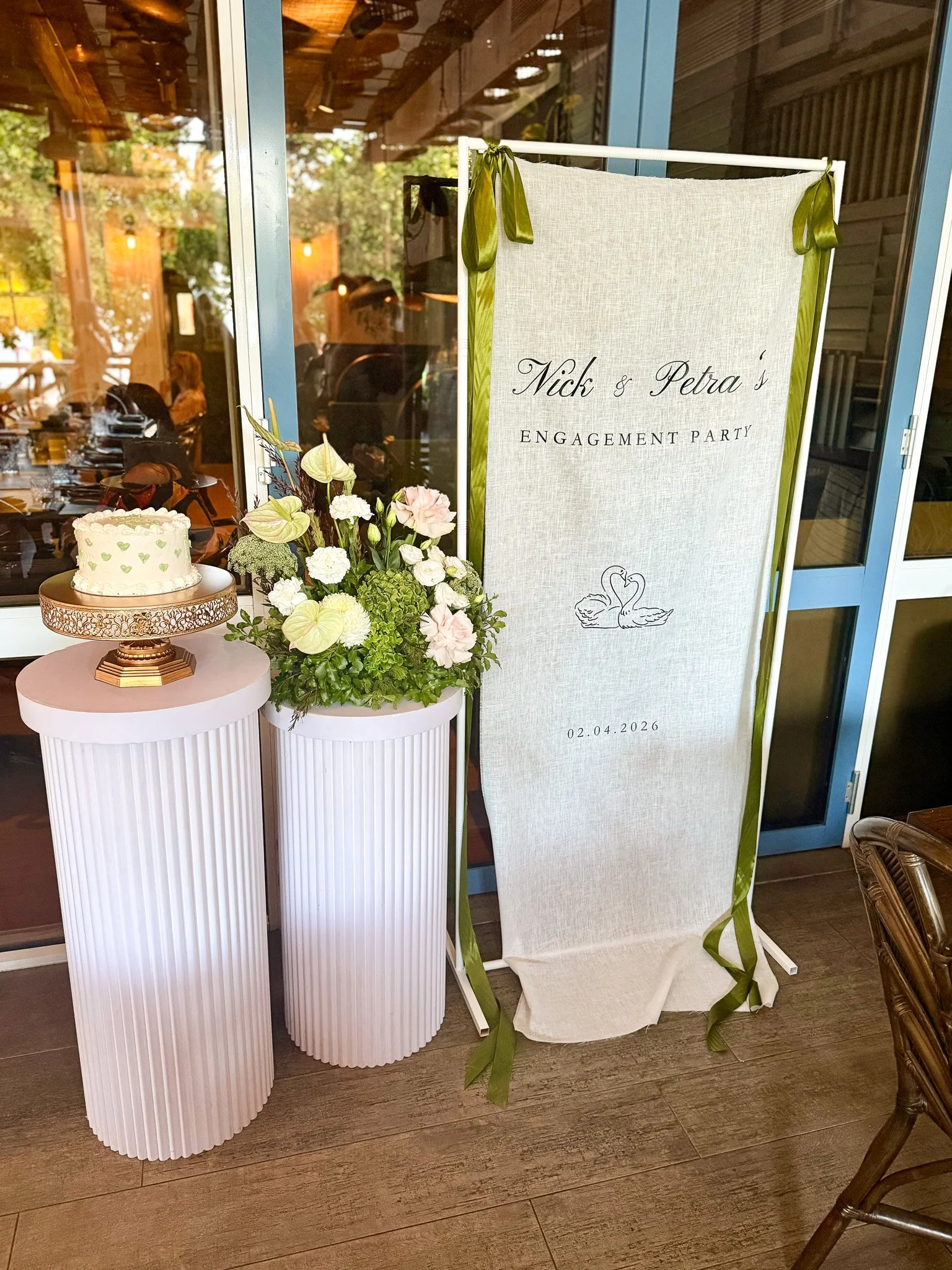A wedding or engagement sign with the names Nick and Petra and the date 02.04.2026, next to a bouquet of flowers and a small cake on a stand, inside a restaurant or event space with glass windows.