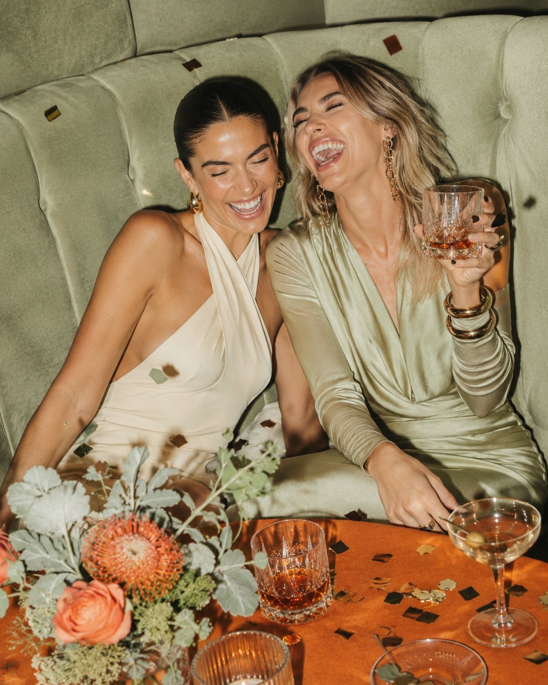 Two women sitting on a green velvet bench at a table, celebrating with drinks and confetti, laughing and enjoying each other's company.