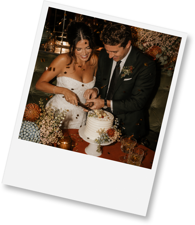 A bride and groom smiling and cutting a wedding cake at their reception, surrounded by flowers and confetti.