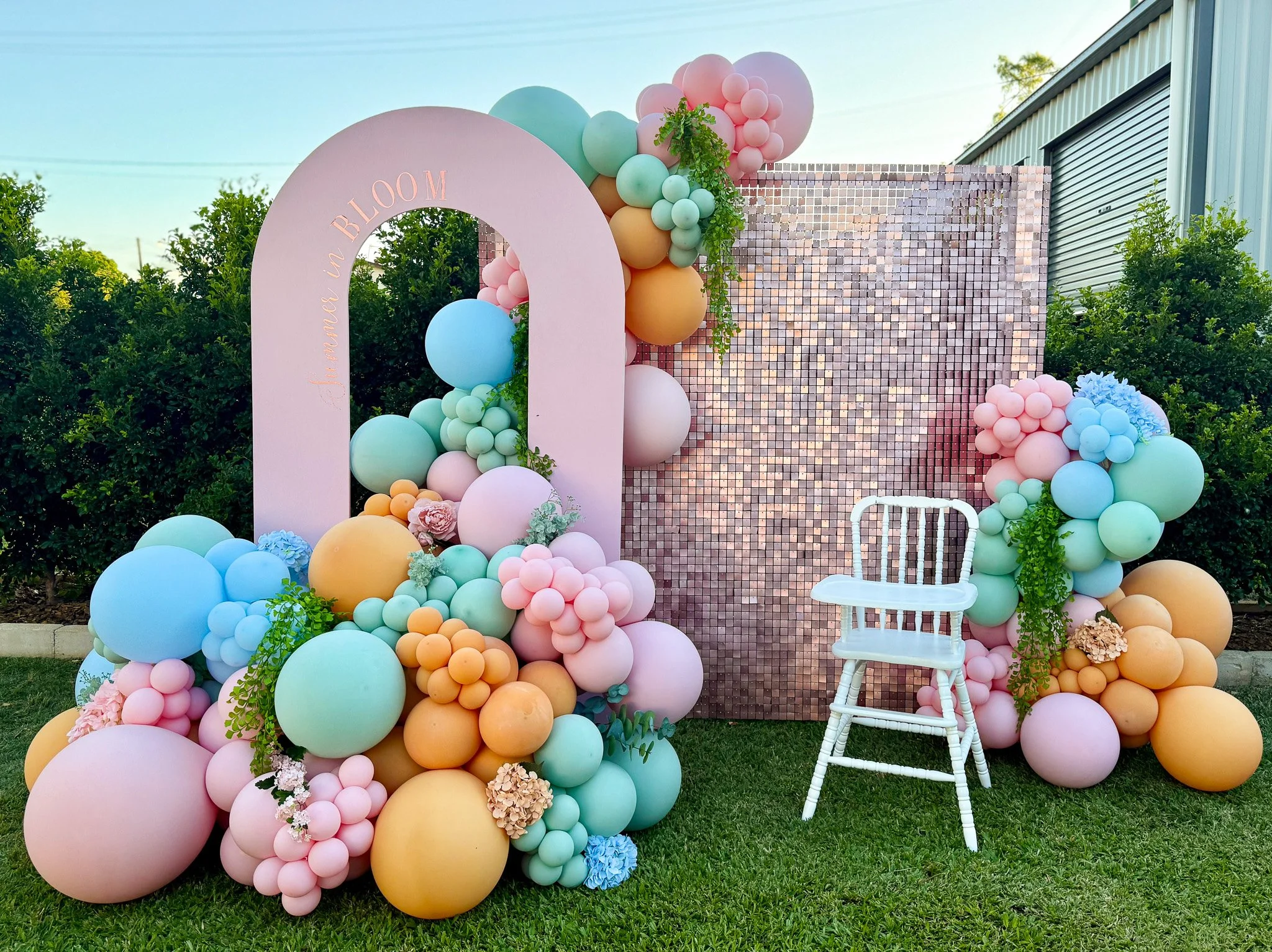 Decorative backdrop with pastel-colored balloons, flowers, and greenery for an outdoor event. Pink, mint, peach, and cream balloons are arranged around a pink arch with the words 'Live in Bloom' written inside, and a small white chair is placed in front.