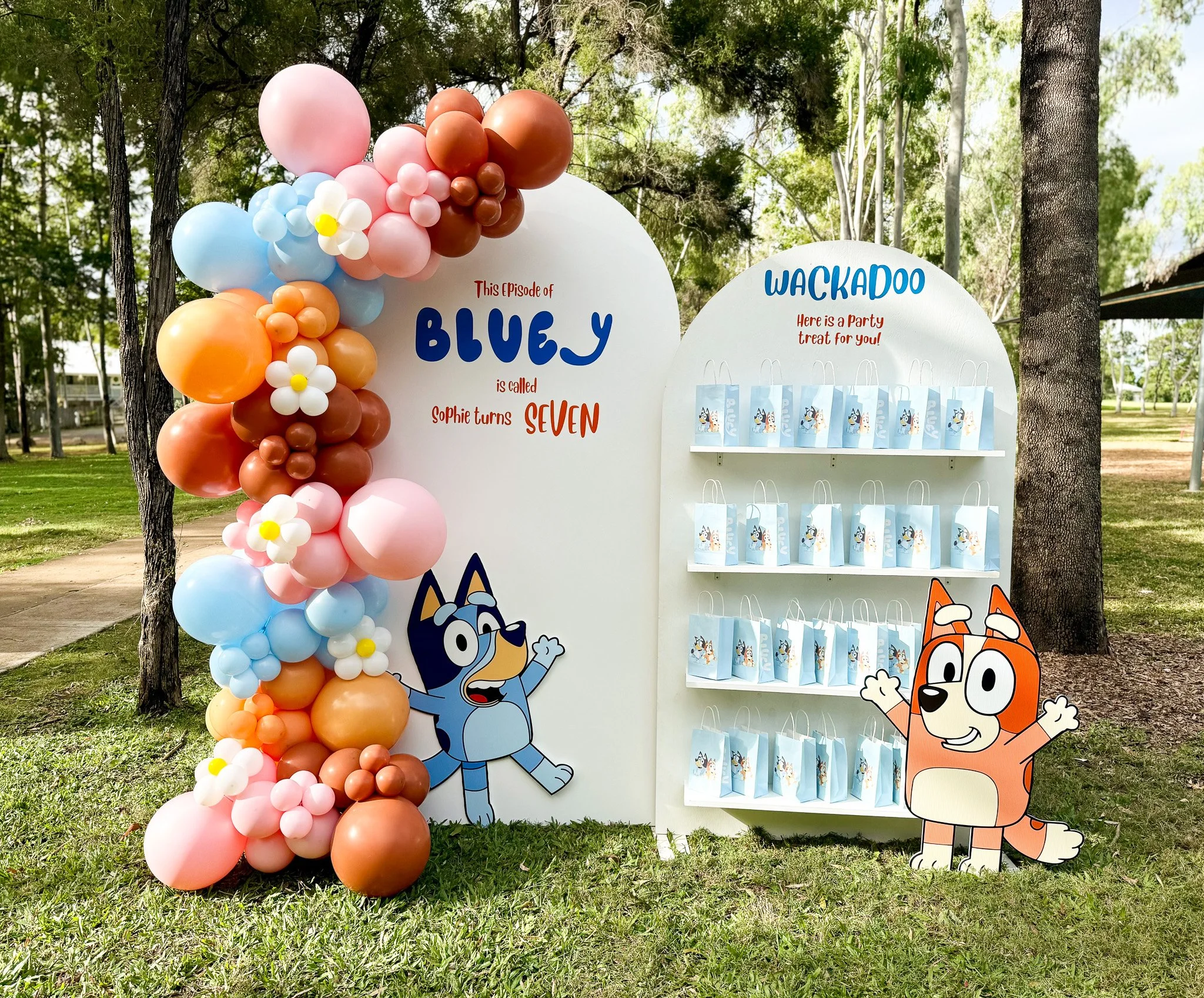 Colorful balloon decoration with pink, orange, blue, and white balloons, including flower-shaped balloons, arranged around a white display board with the text 'This episode of Bluey is called Sophie turns seven' and a separate white display board with party treat bags featuring Bluey characters, set outdoors among trees.