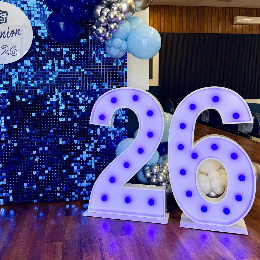 Large illuminated number 20 with purple lights, decorated with balloons in silver, light blue, and dark blue, set against a sparkling blue sequin backdrop at a celebration event.