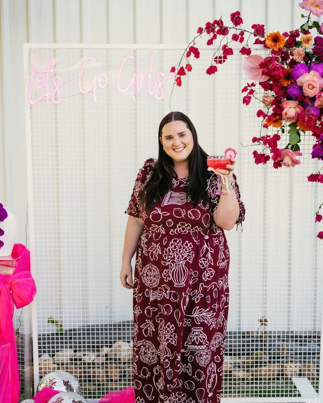 A woman smiling and holding a pink cocktail at a celebration with a 'Let's Go Girls' neon sign, surrounded by pink and purple flowers and decorations.