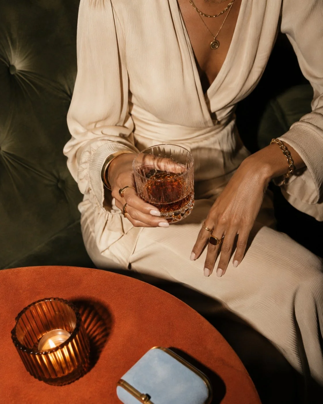 A woman in a cream-colored silky blouse with jewelry holds a glass of dark amber liquid, possibly whiskey or bourbon, while sitting at a table with a lit candle in a decorative holder and a light blue handbag.