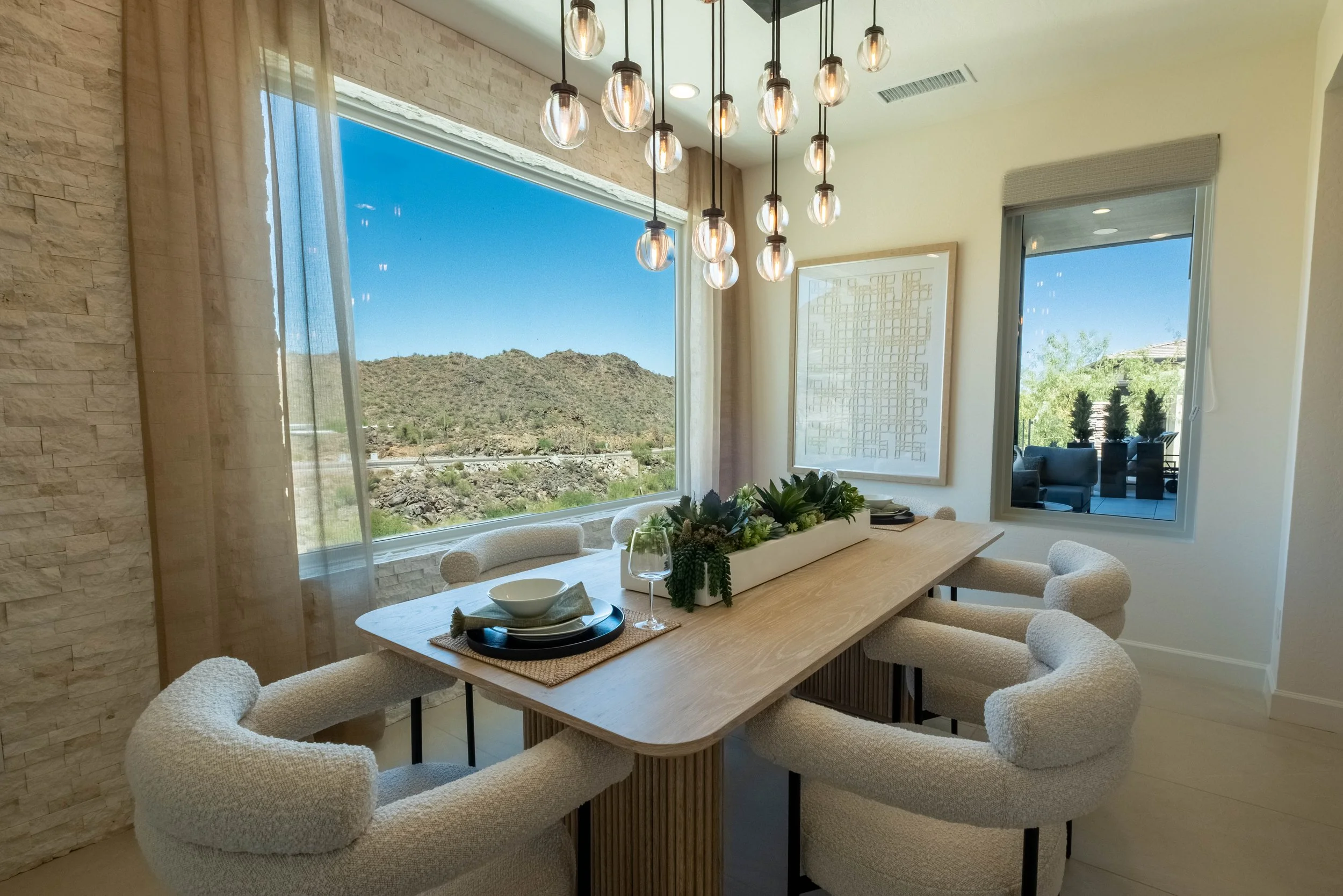 Contemporary dining room with a wooden table, upholstered chairs, and a centerpiece of succulents. Large windows reveal a scenic desert landscape and blue sky. Pendant lights hang above the table.