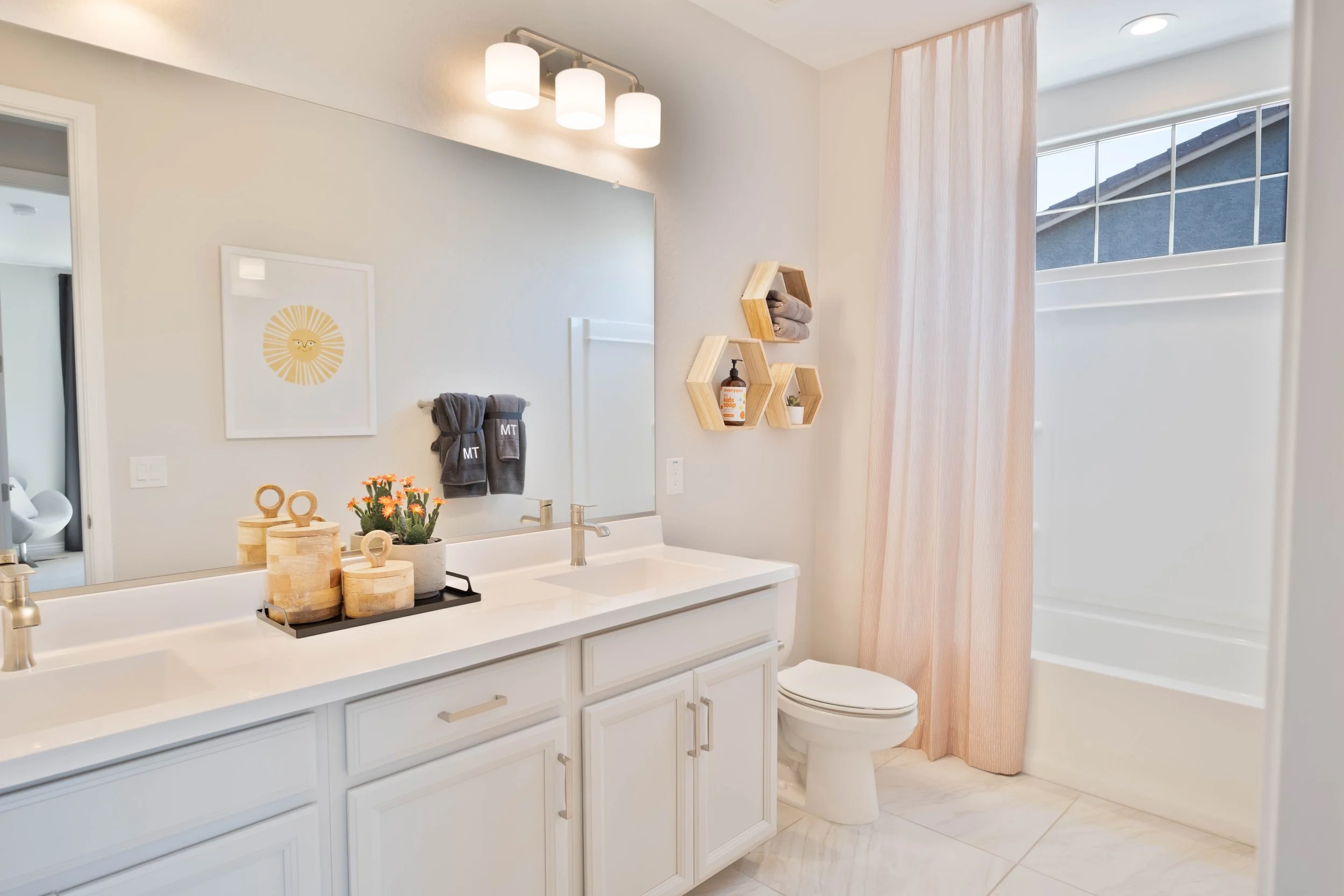 Bright bathroom with white vanity, large mirror, pendant light fixtures, towels, and decorative plants; window with pink curtain and a partial view of the toilet and shower.